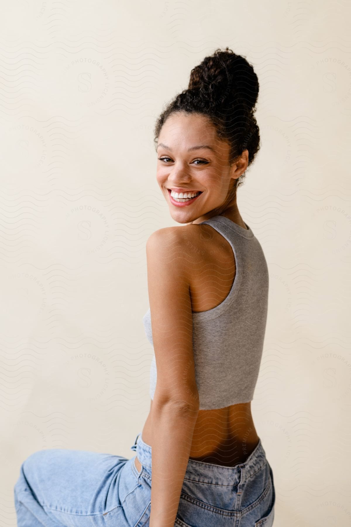 Woman in a gray tank top and blue jeans smiling over her shoulder against a neutral background.