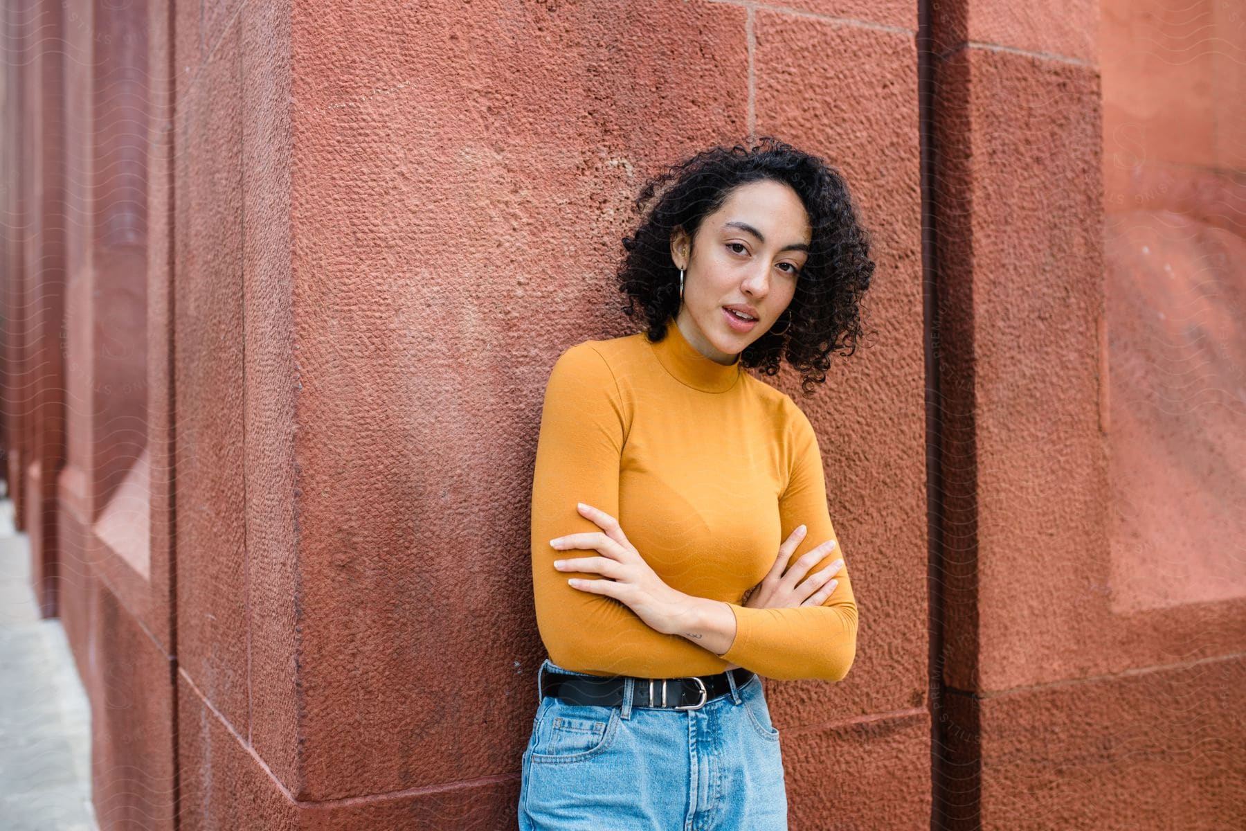 Model leaning against the wall in an orange long-sleeved t-shirt and jeans while arms crossed