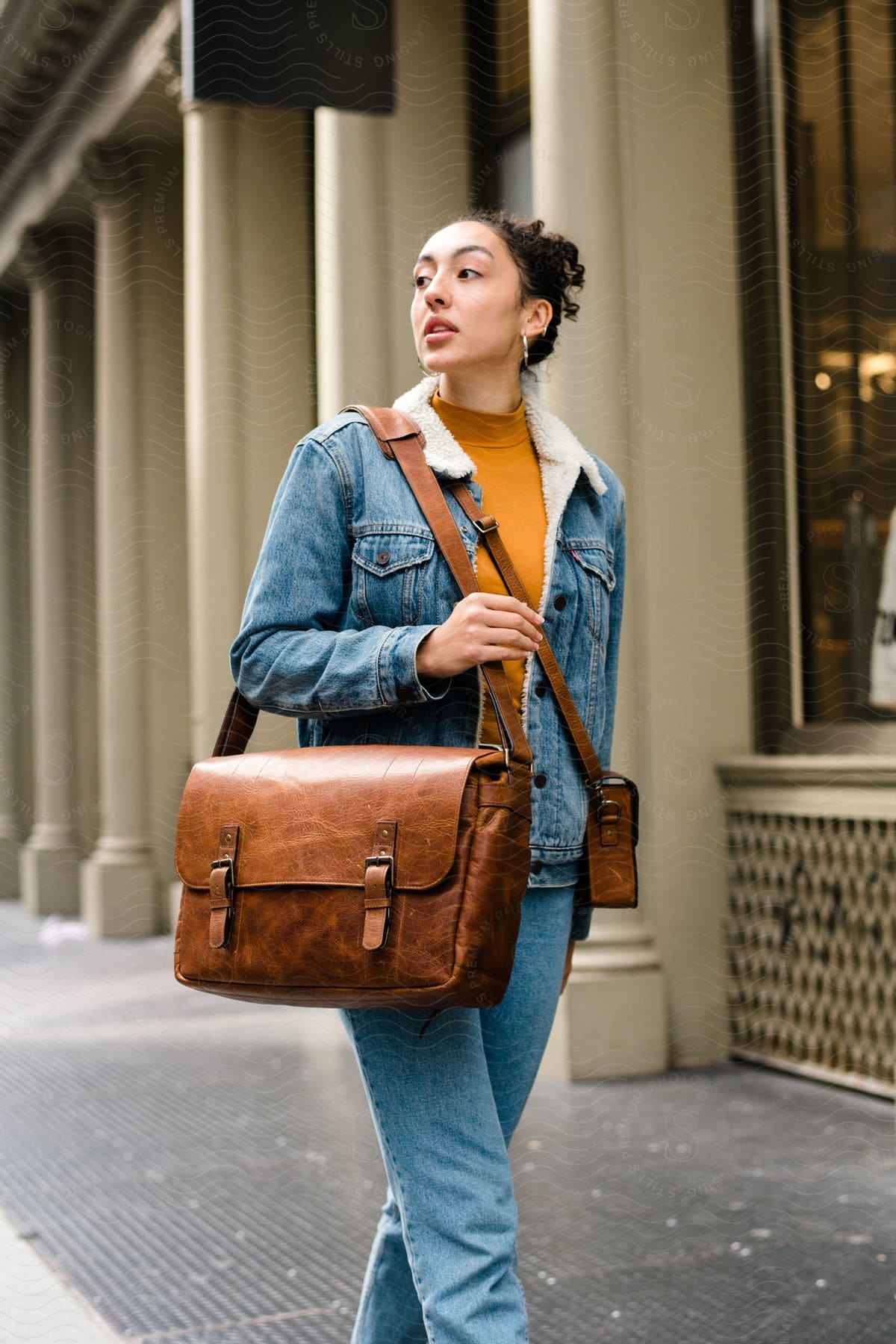 A woman walking outdoors, wearing a denim jacket and carrying a brown leather bag.