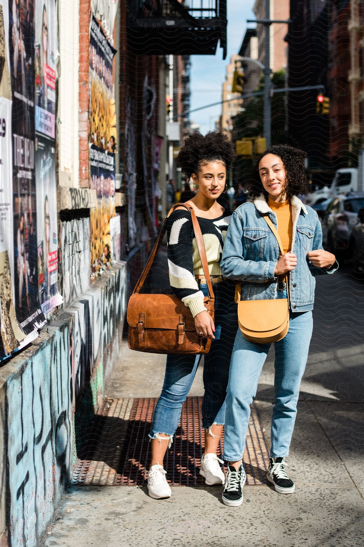 Two women posing on a city sidewalk, one wears a sweater and carries a large brown shoulder bag, the other in a denim jacket with a yellow crossbody bag.