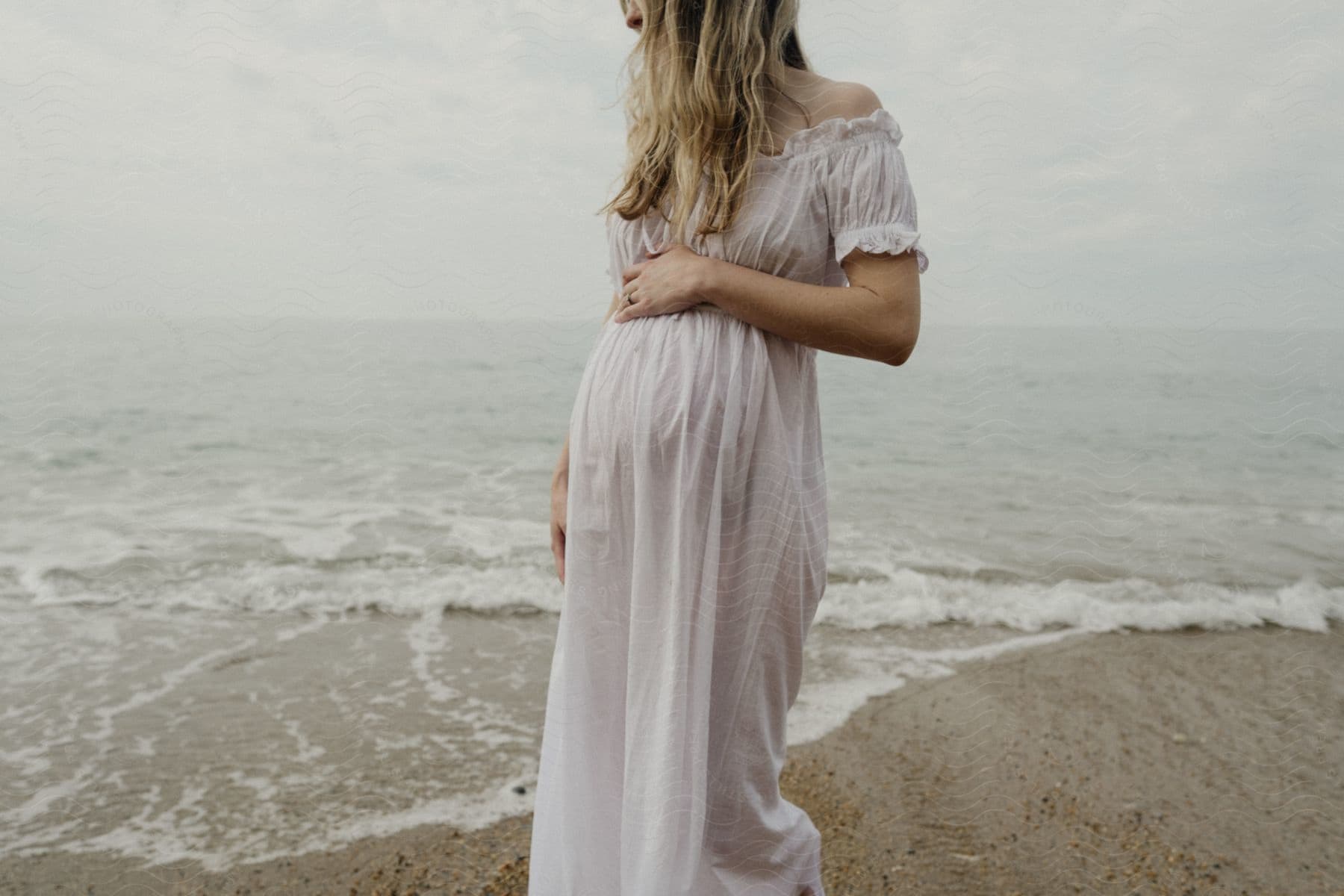 A woman in a flowing dress stands by the sea, holding her belly.