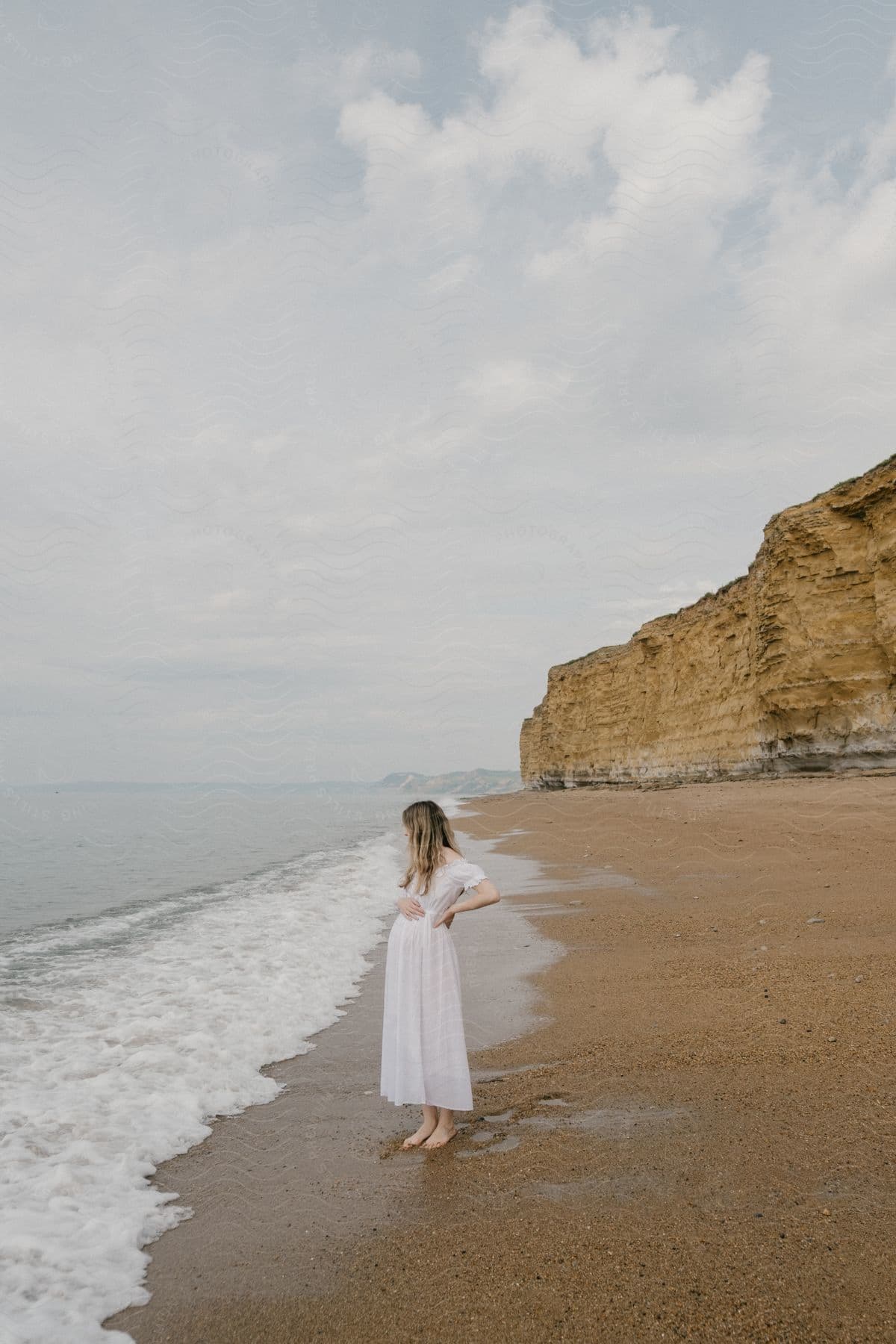 A blonde woman in a white dress stands on a beach with cliffs behind her, waves crashing on shore under cloudy skies.