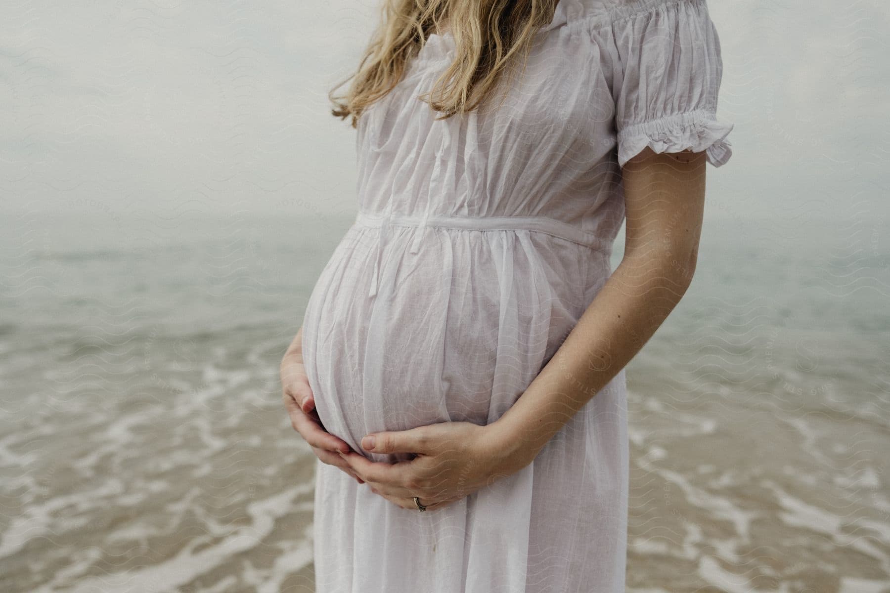 Pregnant woman in a white dress holding her belly by the sea.