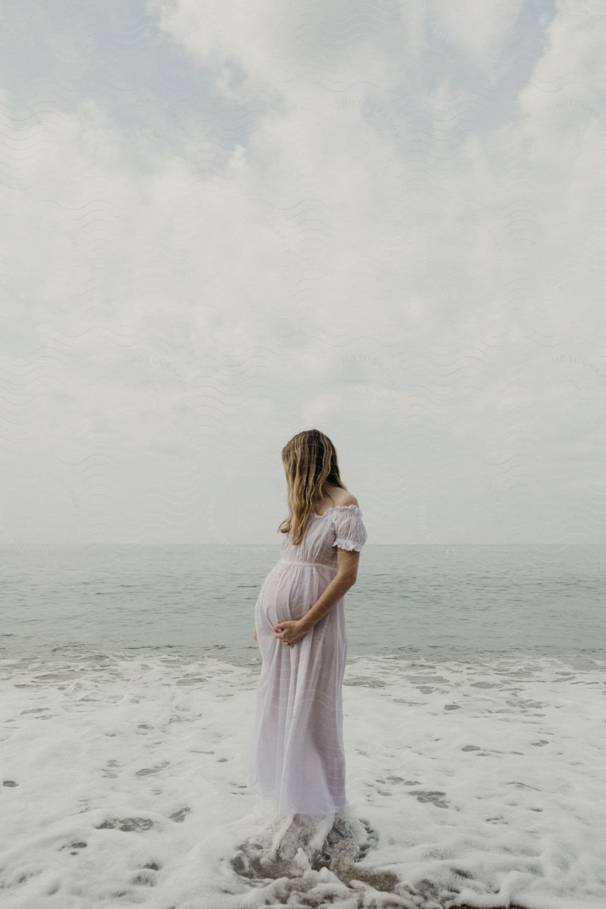 Pregnant woman in a white dress standing by the sea.