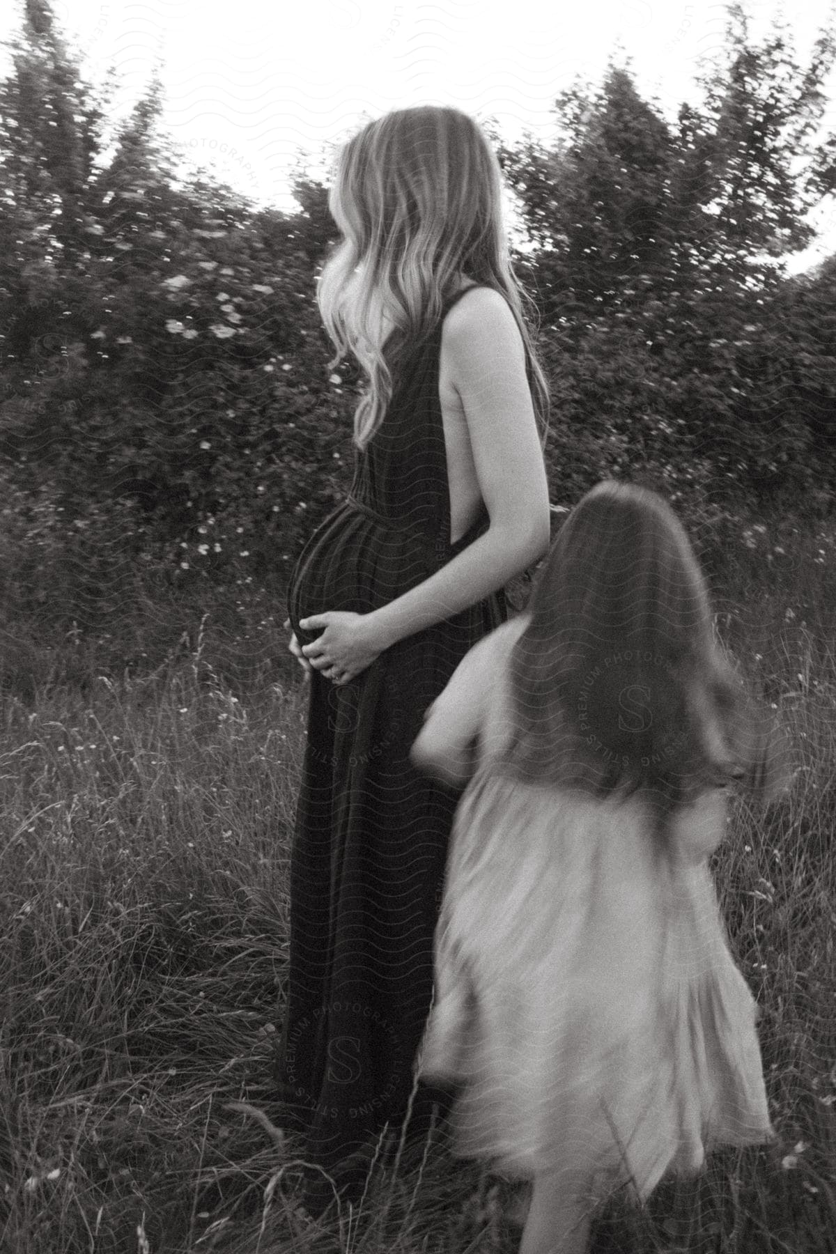 Girl Walks Around Pregnant Woman Standing In Field Of Grass