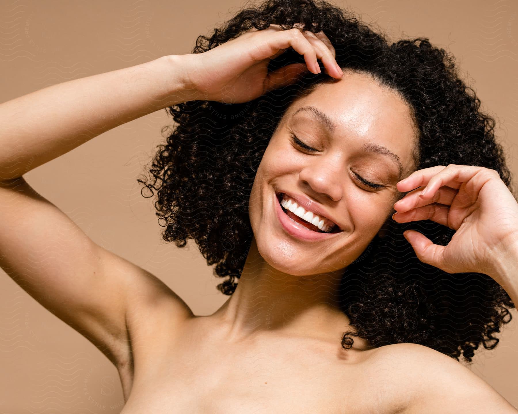 Joyful woman with curly hair stretching with hands raised and eyes closed, bare shoulders visible, against a beige background.