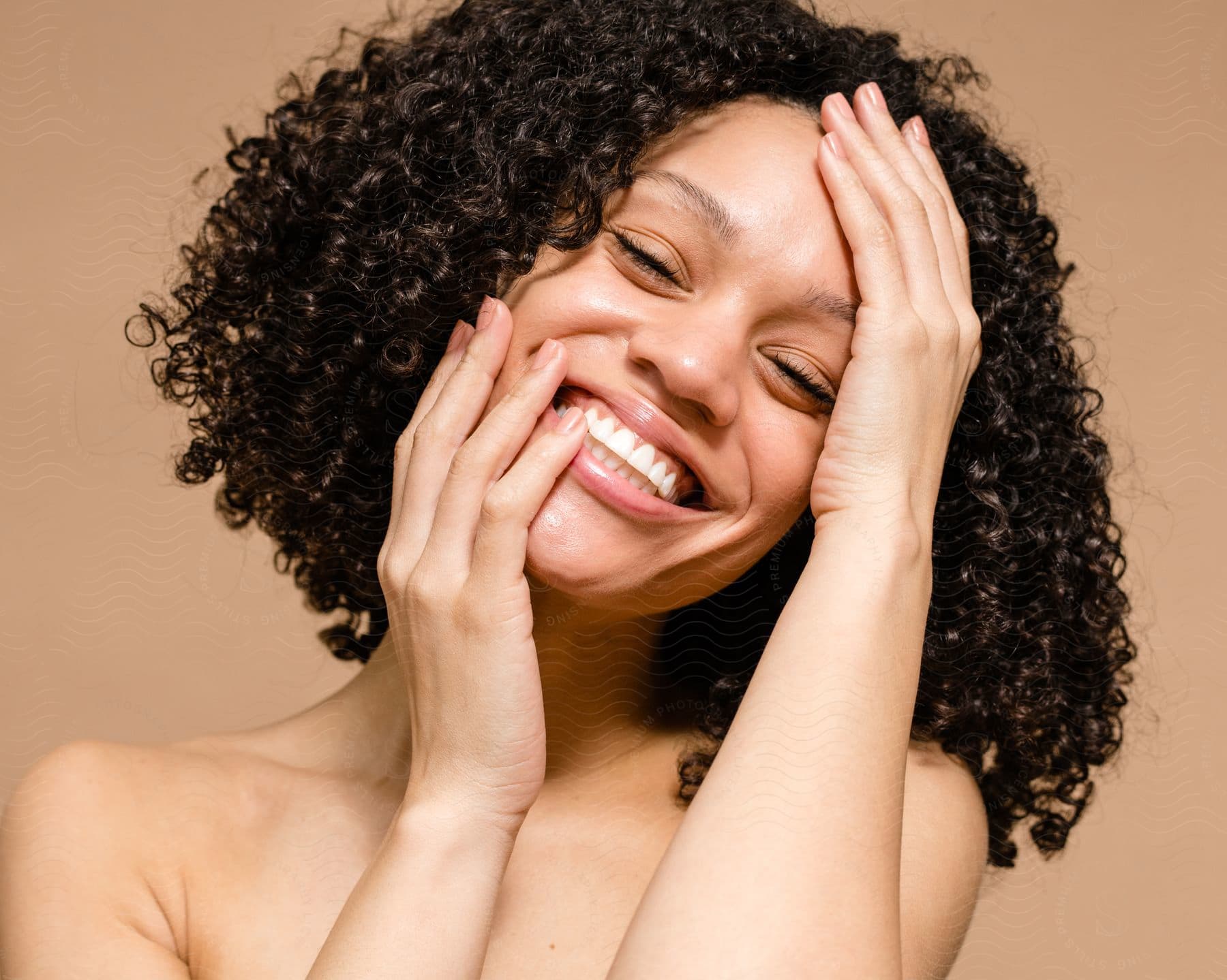 Portrait of a woman with curly black hair and smiling with her hands on her face.