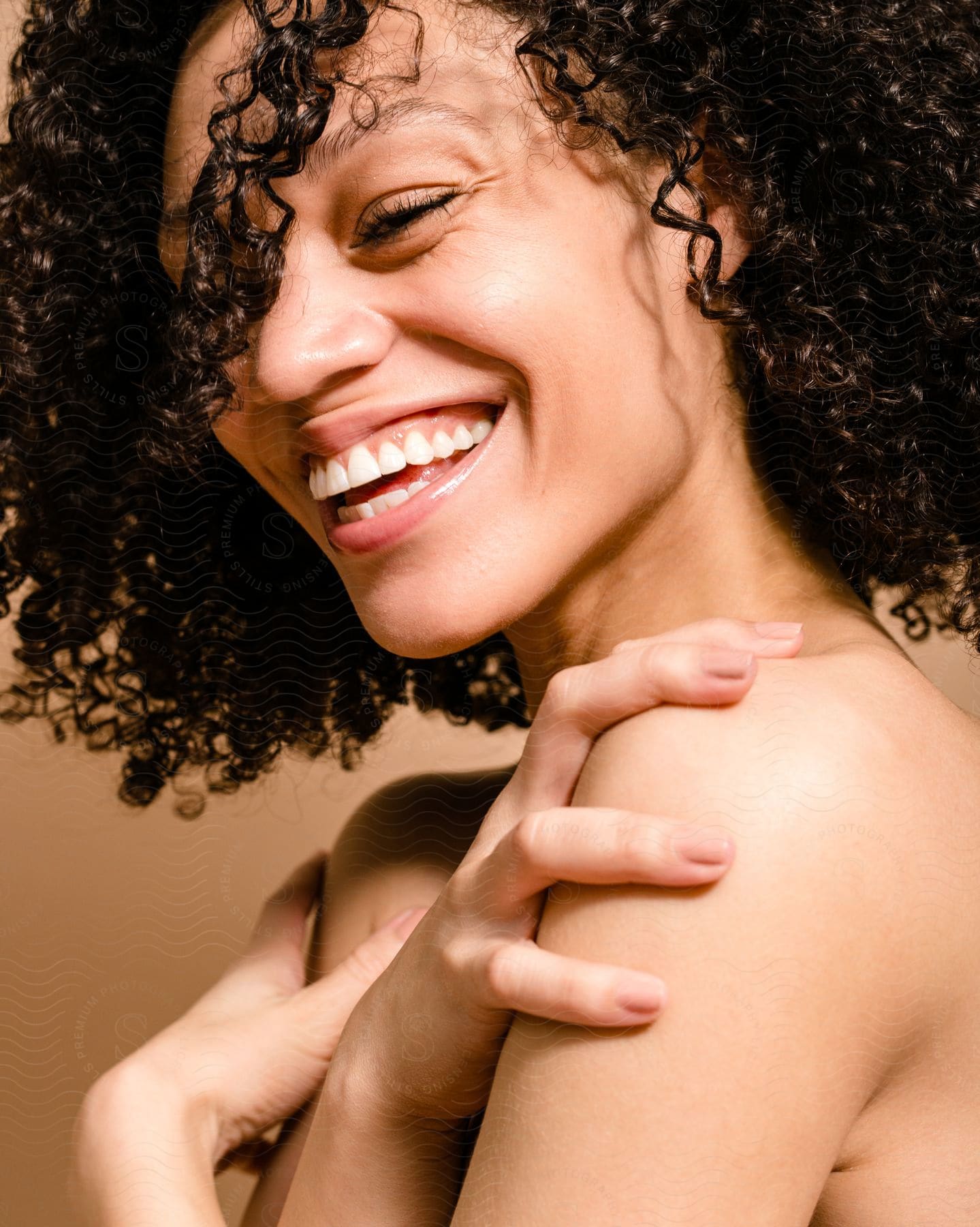 A topless woman with curly hair laughs while holding her shoulders.