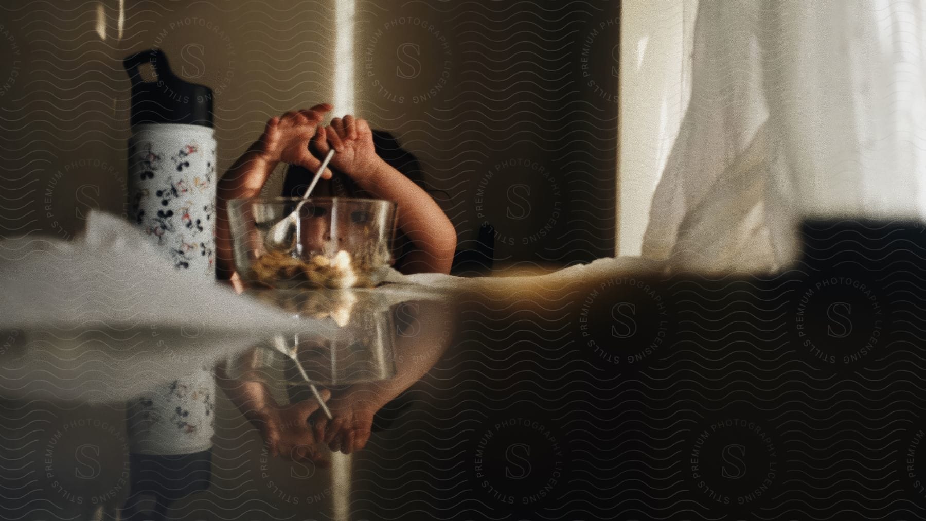 Little girl sitting at a table next to a window with a white curtain, holding a spoon in a glass bowl with food.