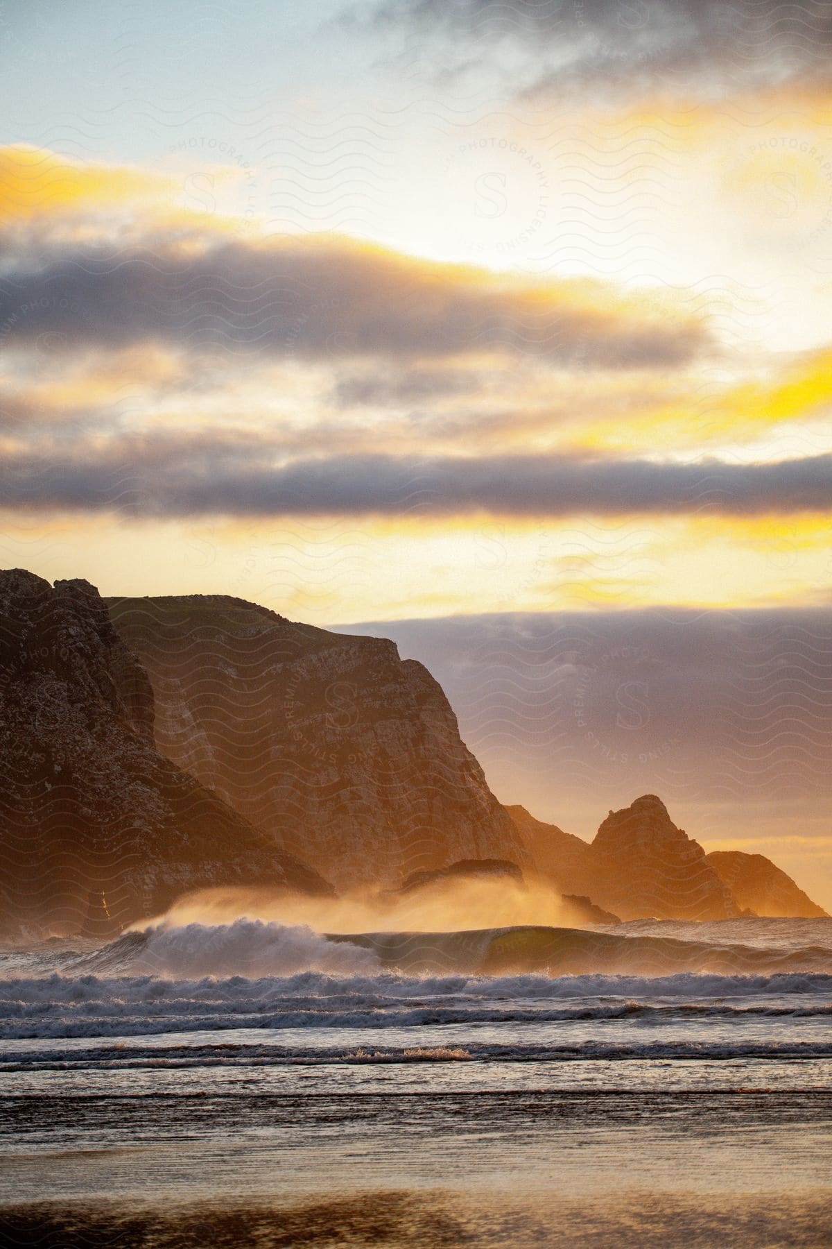 A serene sunset scene illuminates the hazy waves crashing against the rocky cliffs.