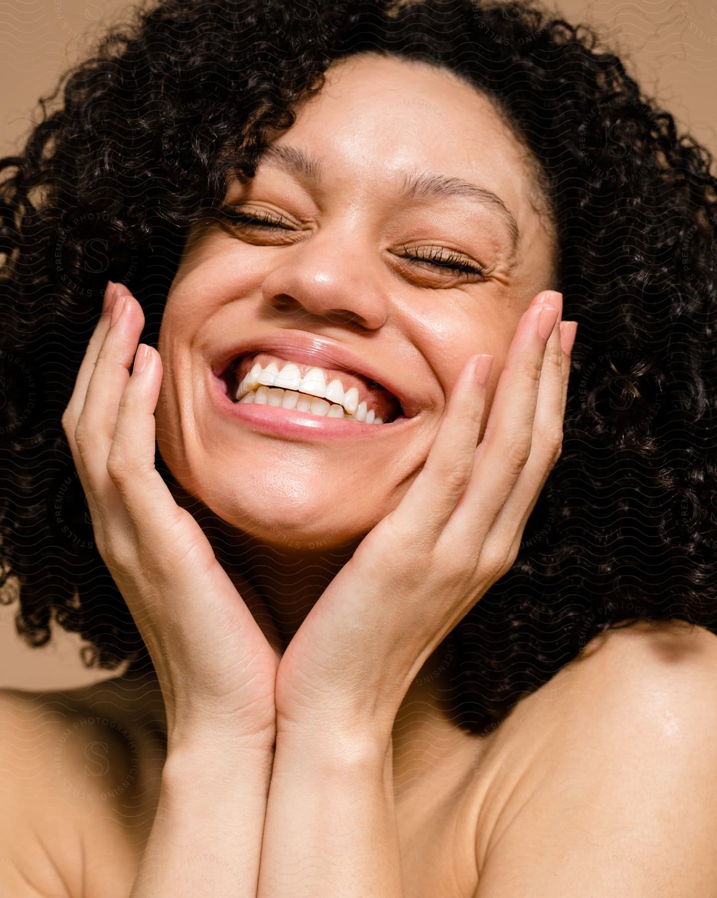 Portrait of a young woman with black curly hair with her eyes closed smiling and with her two palms on her face