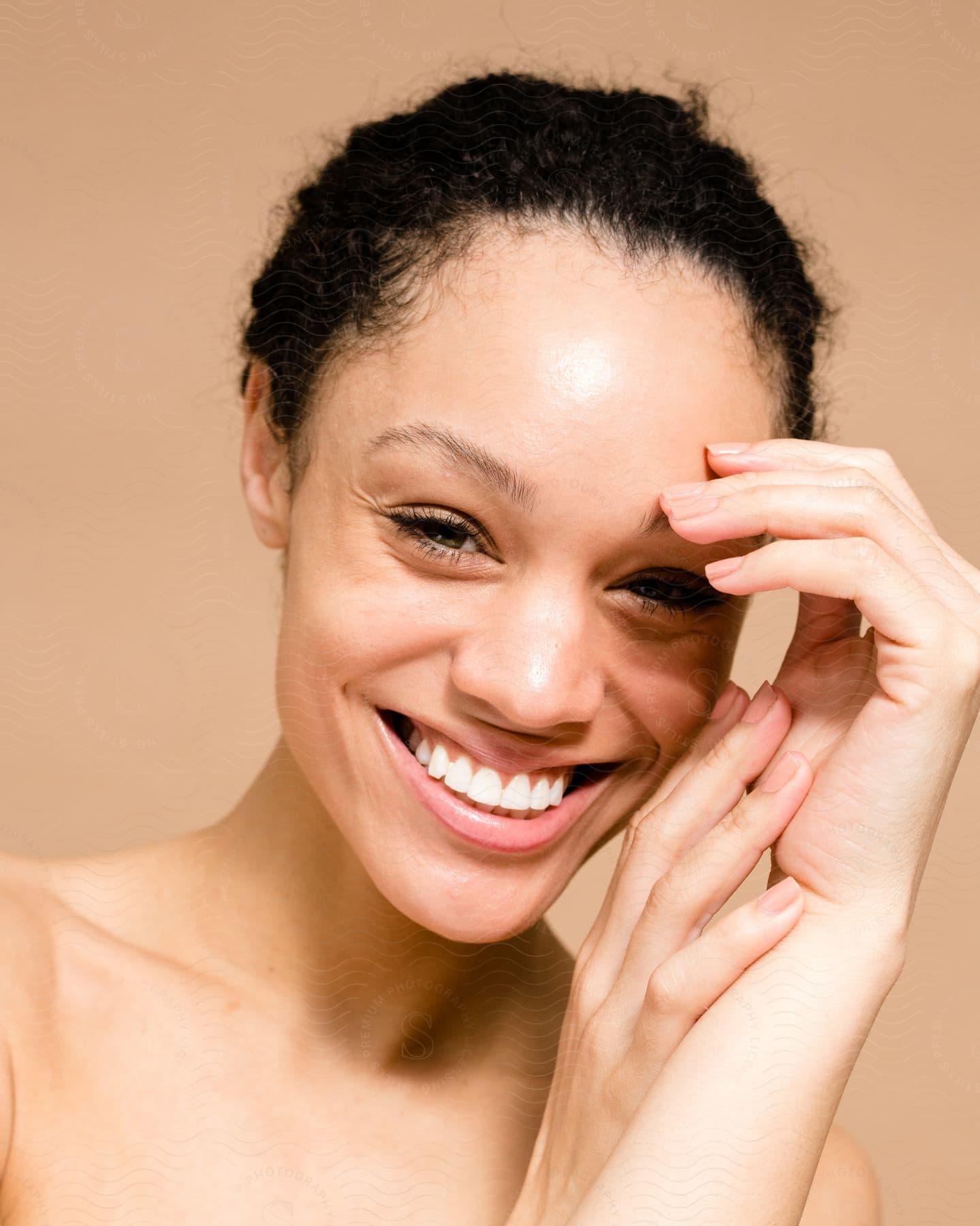 A young woman is smiling while holding her two hands and the left part of her face and she is looking straight ahead