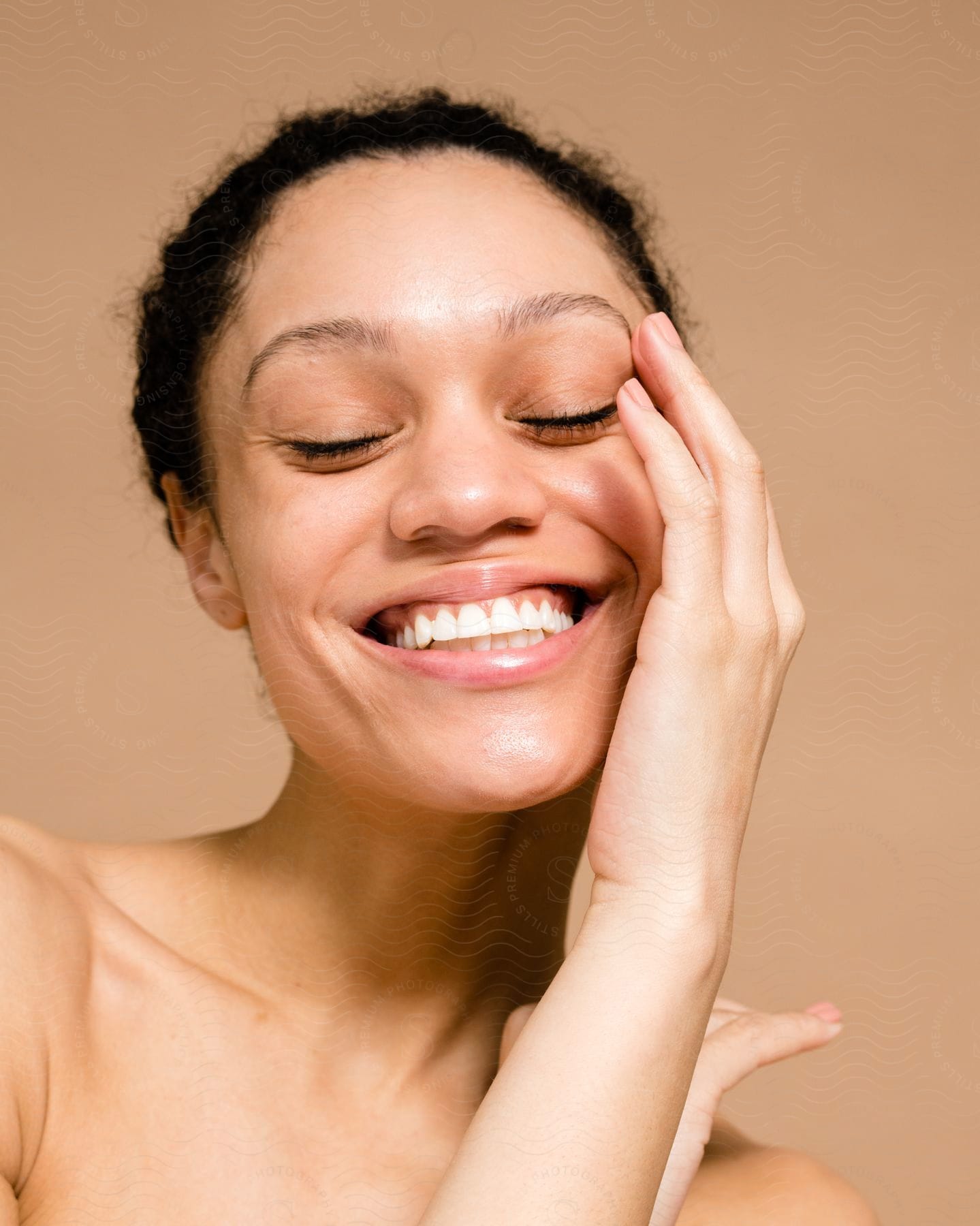 Portrait of a woman with her eyes closed, her hair tied up and smiling with one hand on her face.