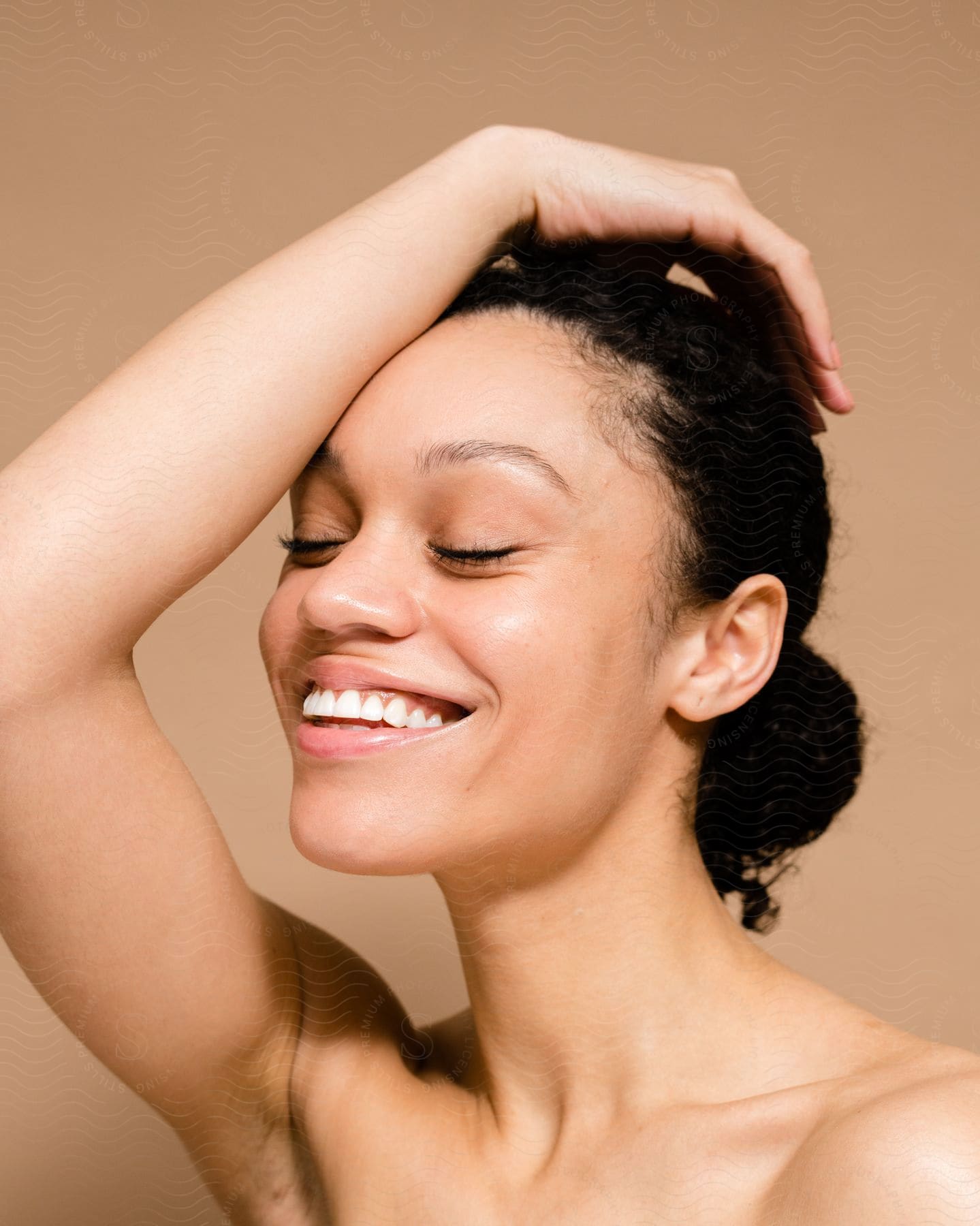 Close-up on a woman's face with her black hair tied up and one of her hands on her head while smiling