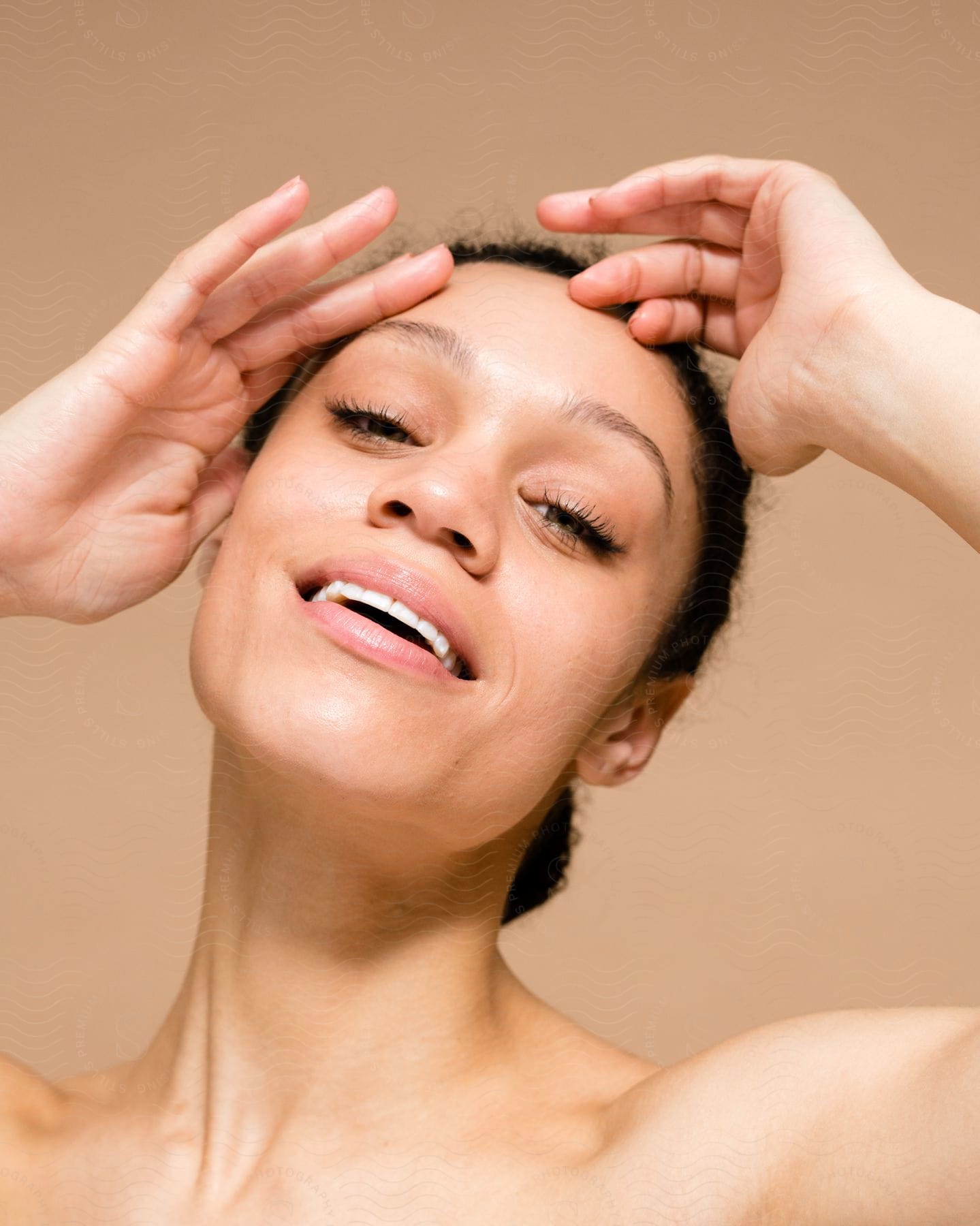 Portrait of a young woman with black hair tied up, giving a slight smile and with her palms under her face on a background contrasting with skin color and blurred.