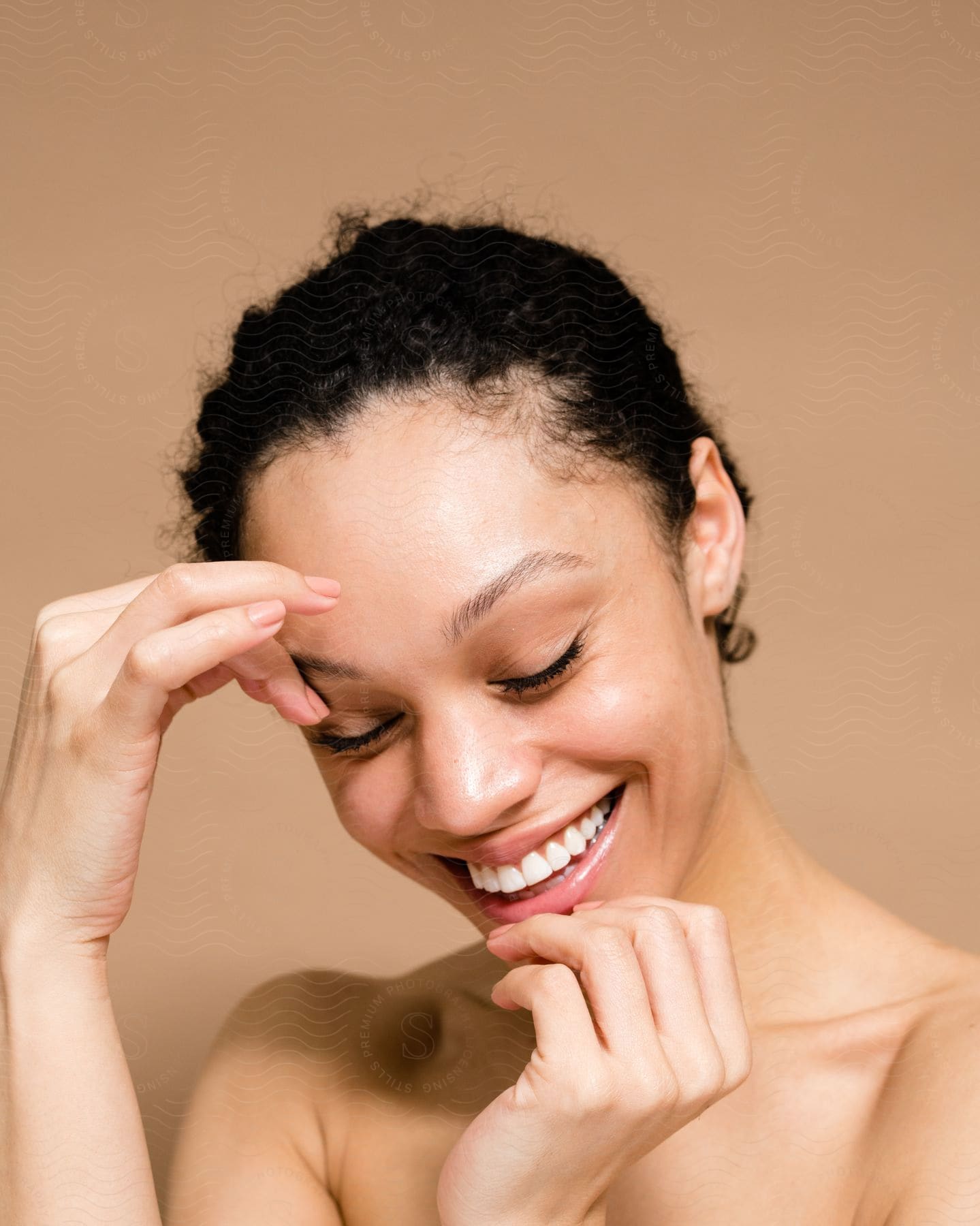 A topless woman with curly hair touches her jaw and eyebrow while laughing.