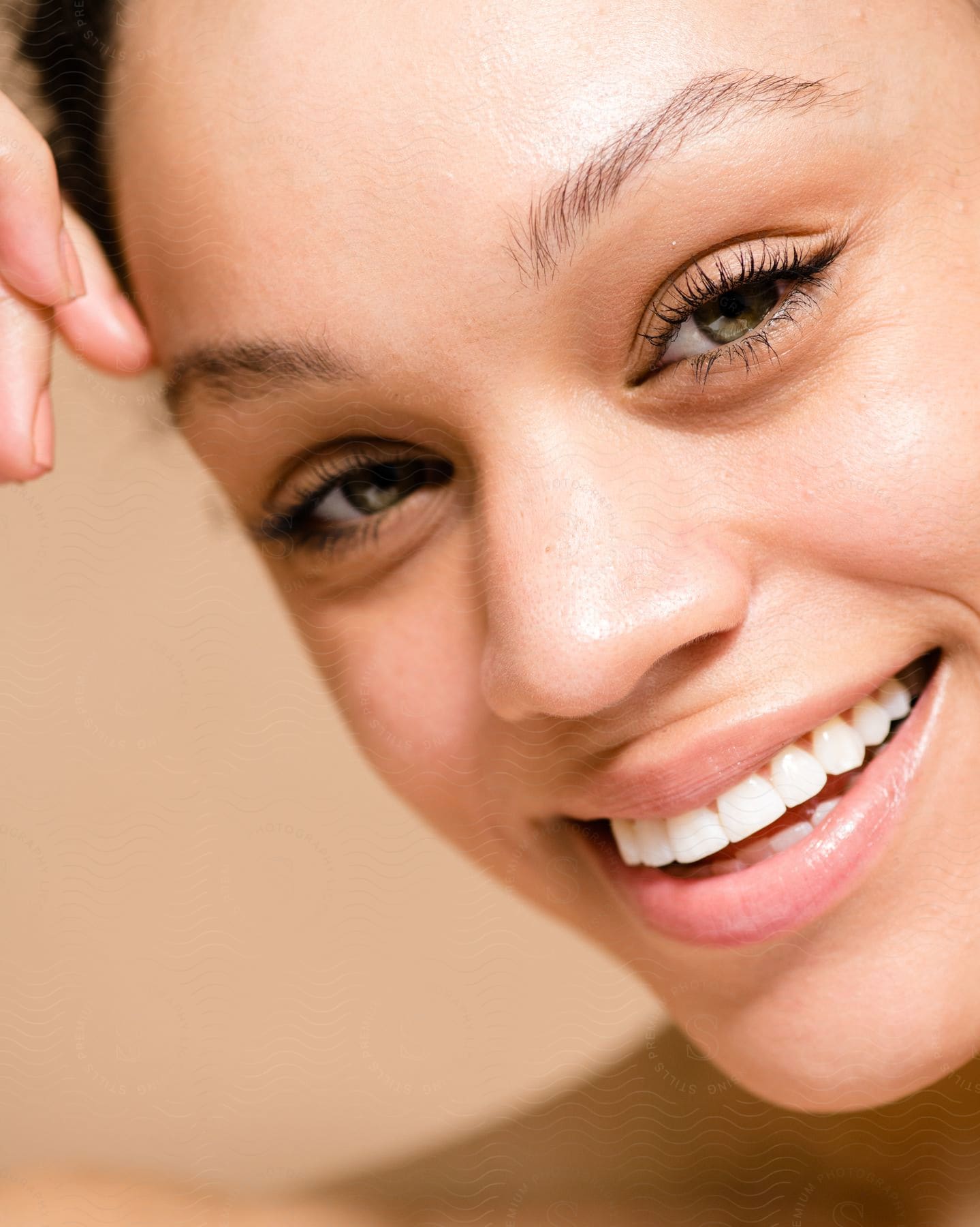 The face of a woman who is smiling and showing her white teeth and green eyes while her fingers touch her forehead