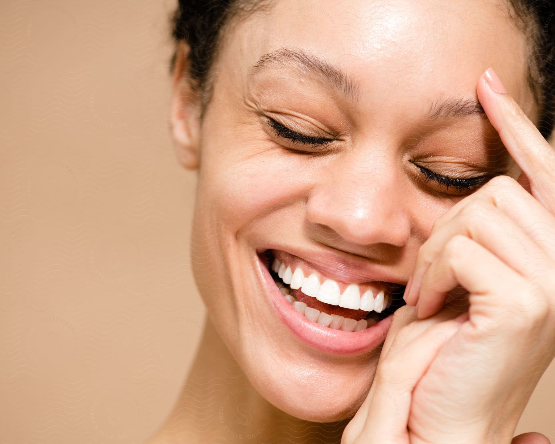 Laughing woman with eyes closed and hand on forehead against a beige background.