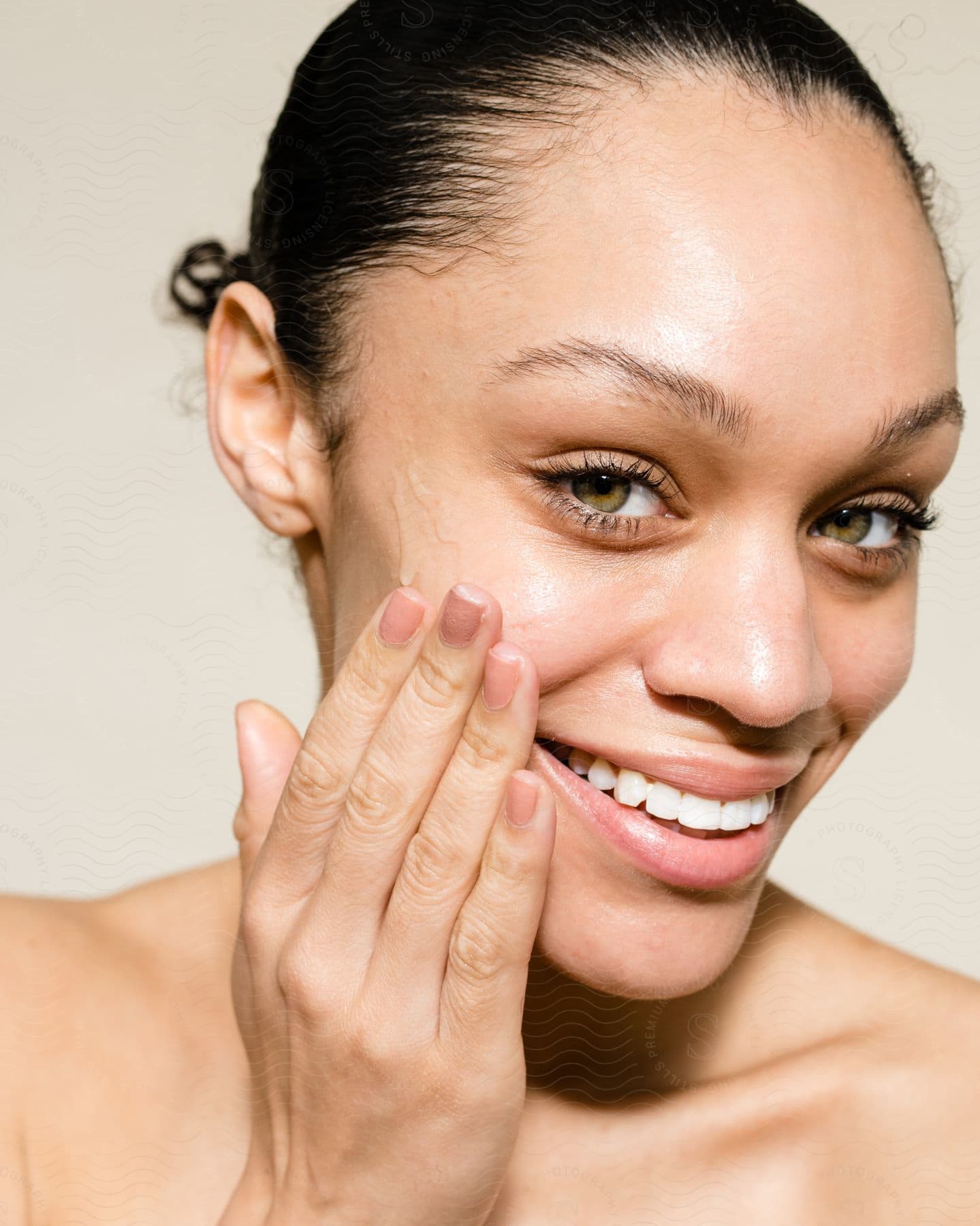 Young smiling woman with her hair tied up applying skincare liquid with the help of her hand on her face.