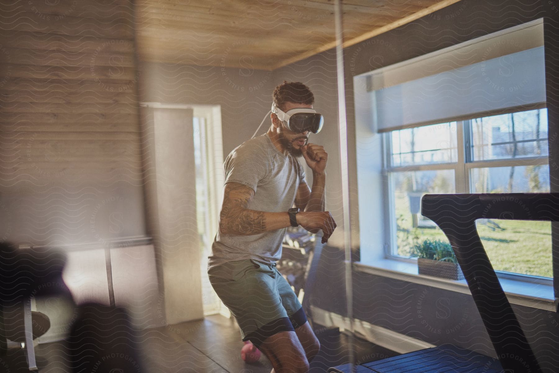 A tattooed man wearing a VR headset is jogging in place inside a room with a treadmill and window.