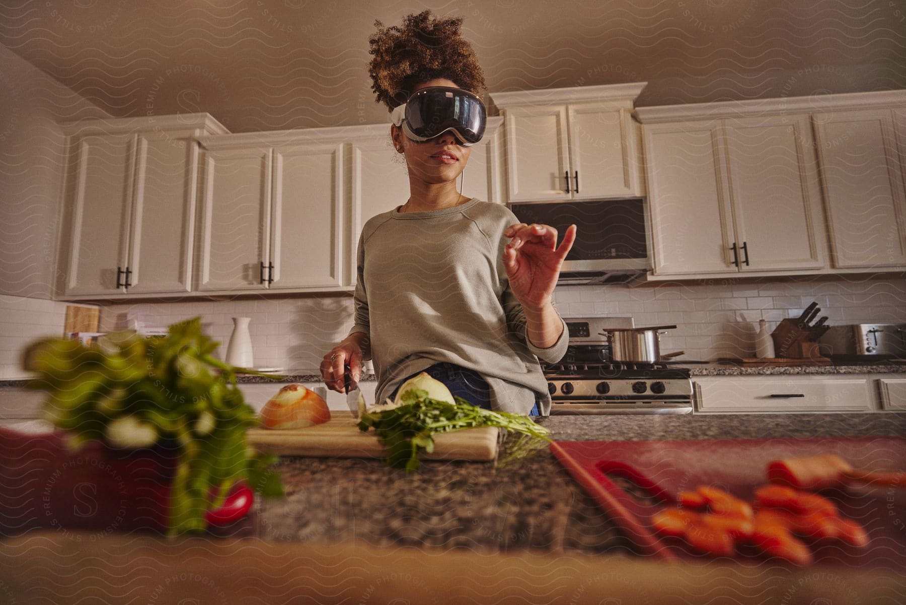 A womanwearing a large pair of VR goggles gestures in a kitchen with vegetables and a cutting board in front of her.