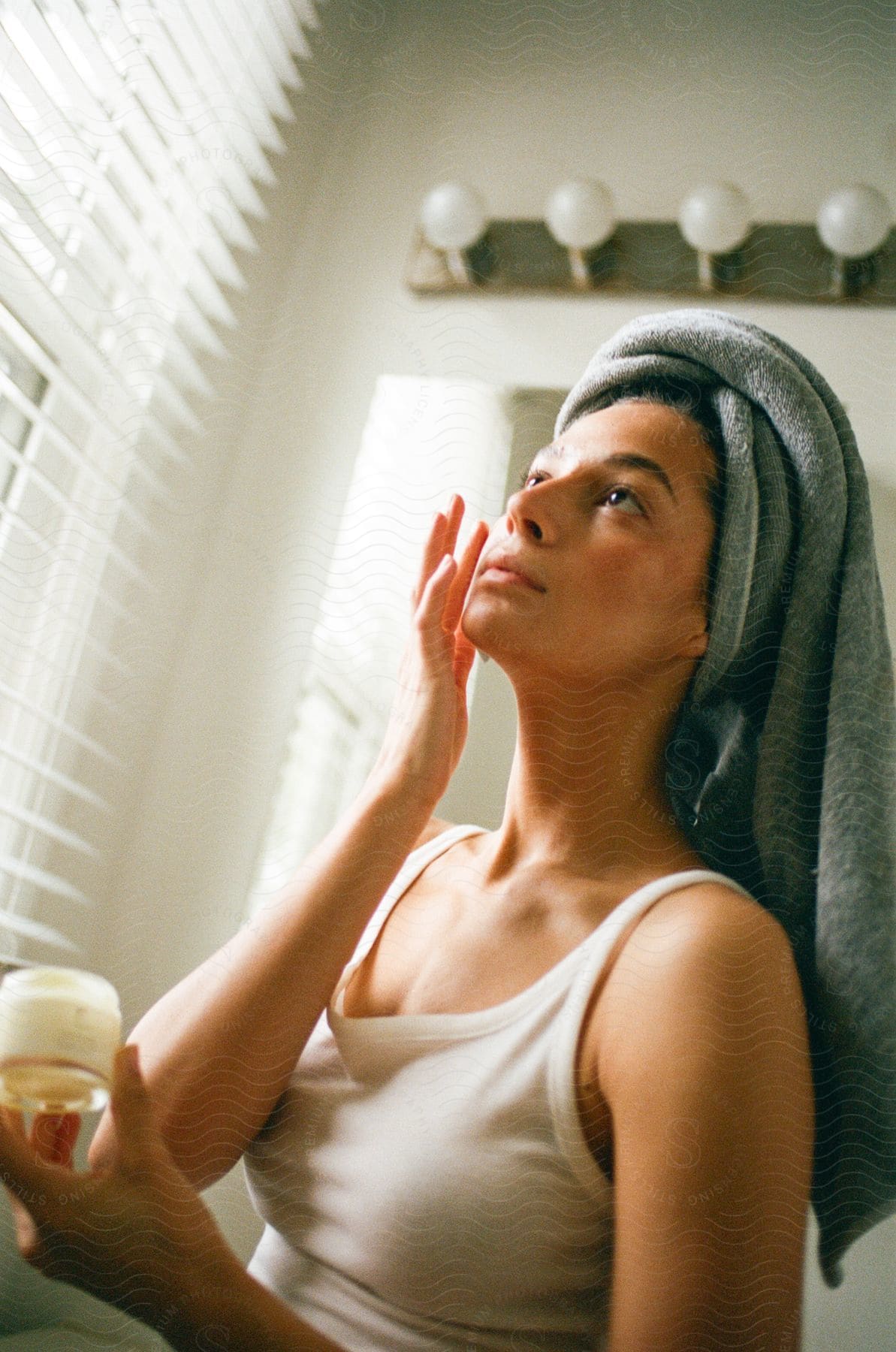 A woman with a towel wrapped around her head applies facial cream while looking upwards, standing in a bathroom with blinds.