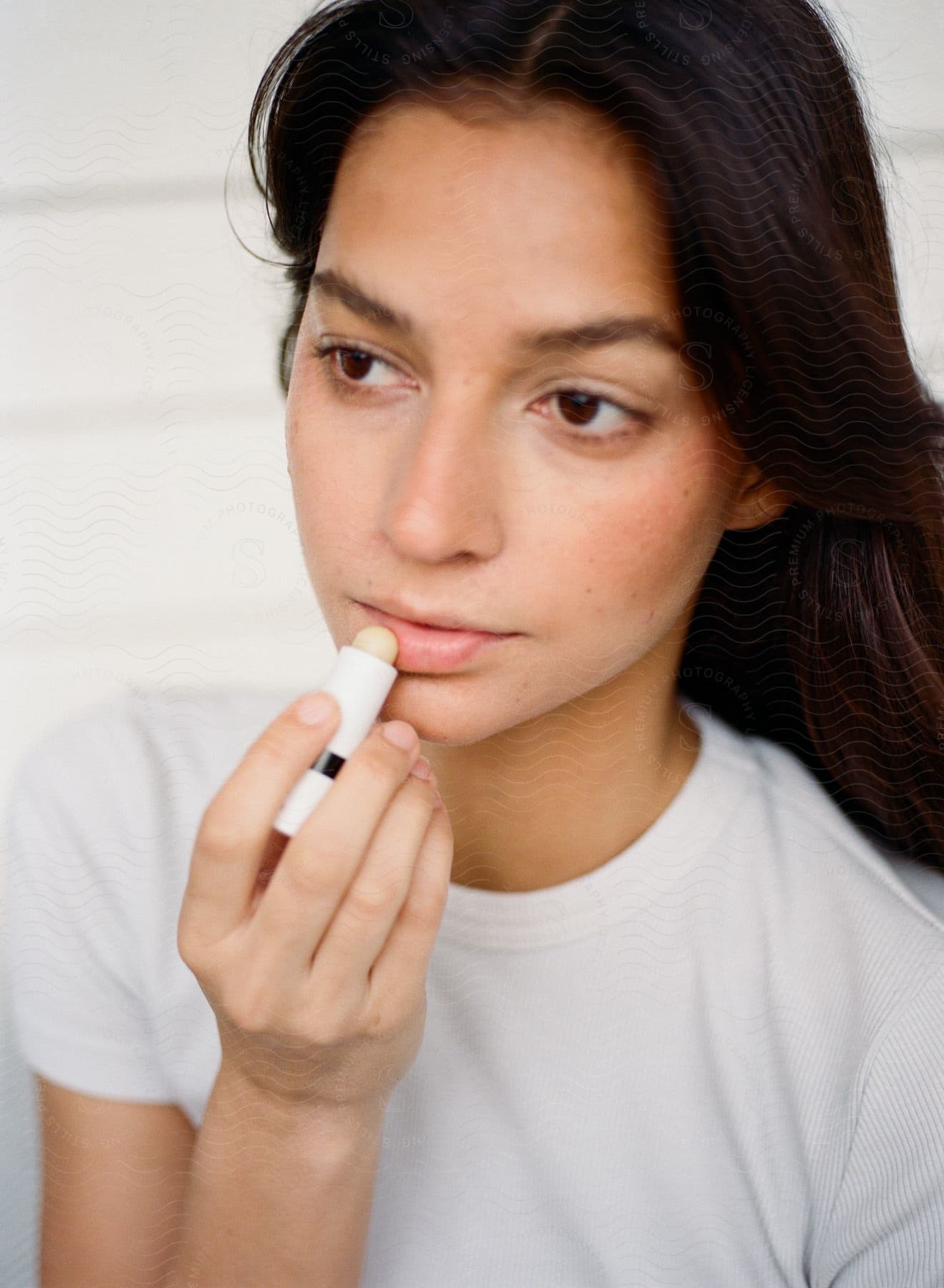 A woman in a white shirt applies lip balm while gazing thoughtfully to the side.