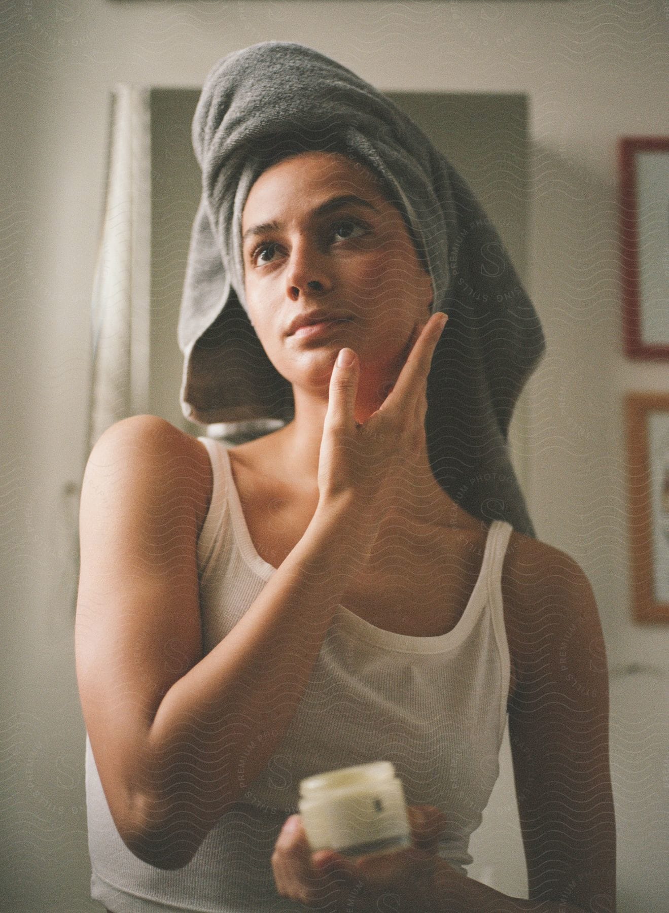 A woman in a tank top and a towel around her head applies skin cream to her face.