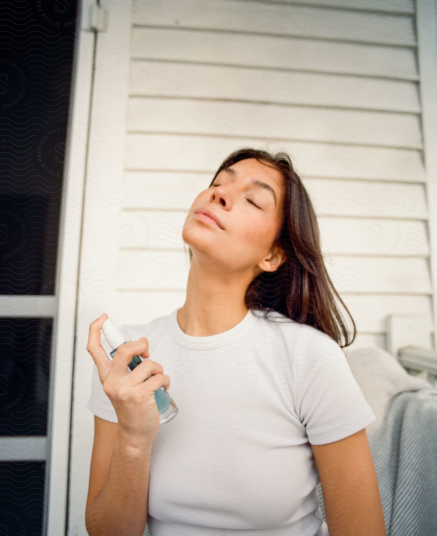 Woman with eyes closed, spraying a bottle towards her face, seated against a white backdrop.