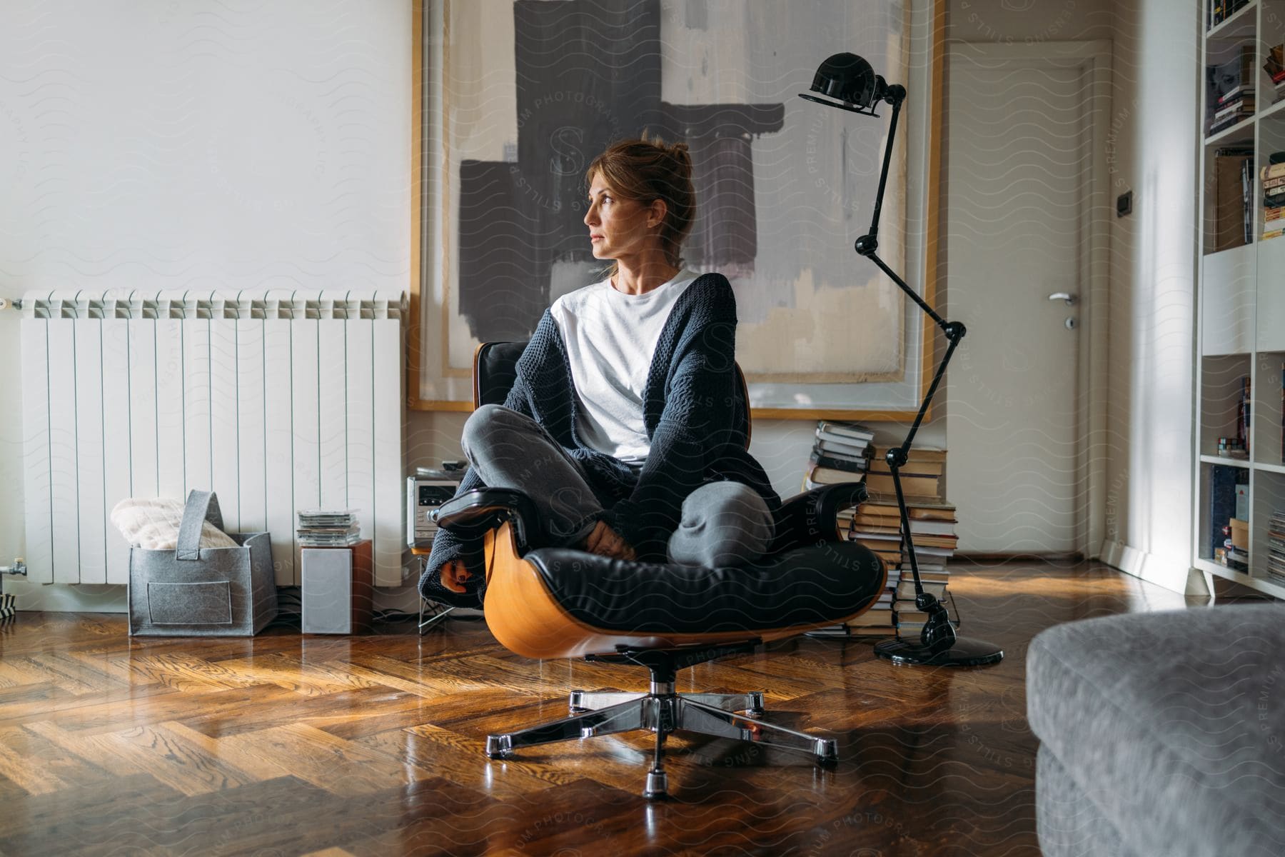 A woman posed sitting in a modern desk chair near a jointed floor lamp books and an abstract painting