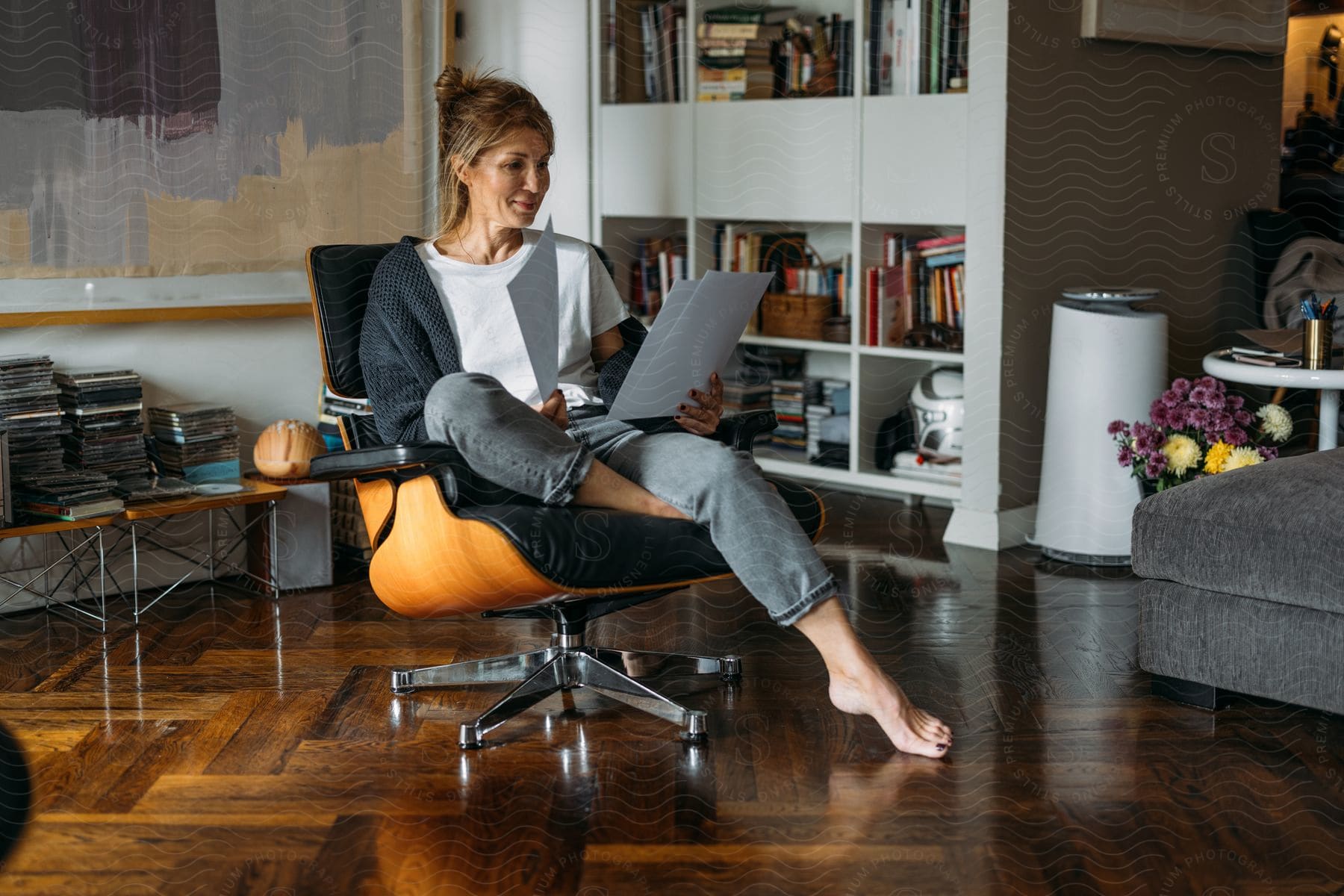 Woman sitting in a leather armchair reading documents while in a room with wooden laminate flooring and a white bookshelf in the background with books.