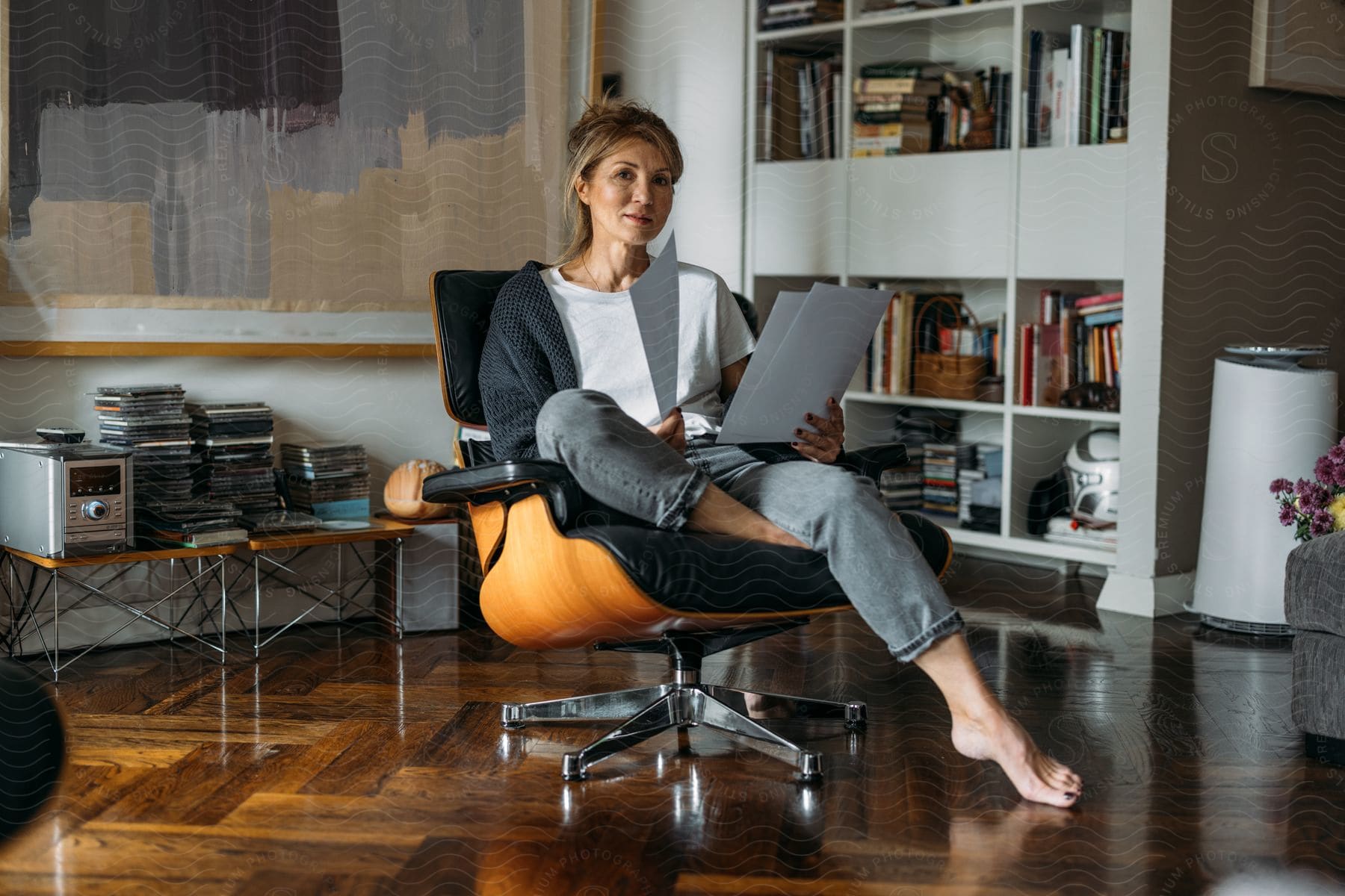 Woman sitting in an armchair with documents in her hand, in a well-lit and organized home environment, with a bookshelf in the background.