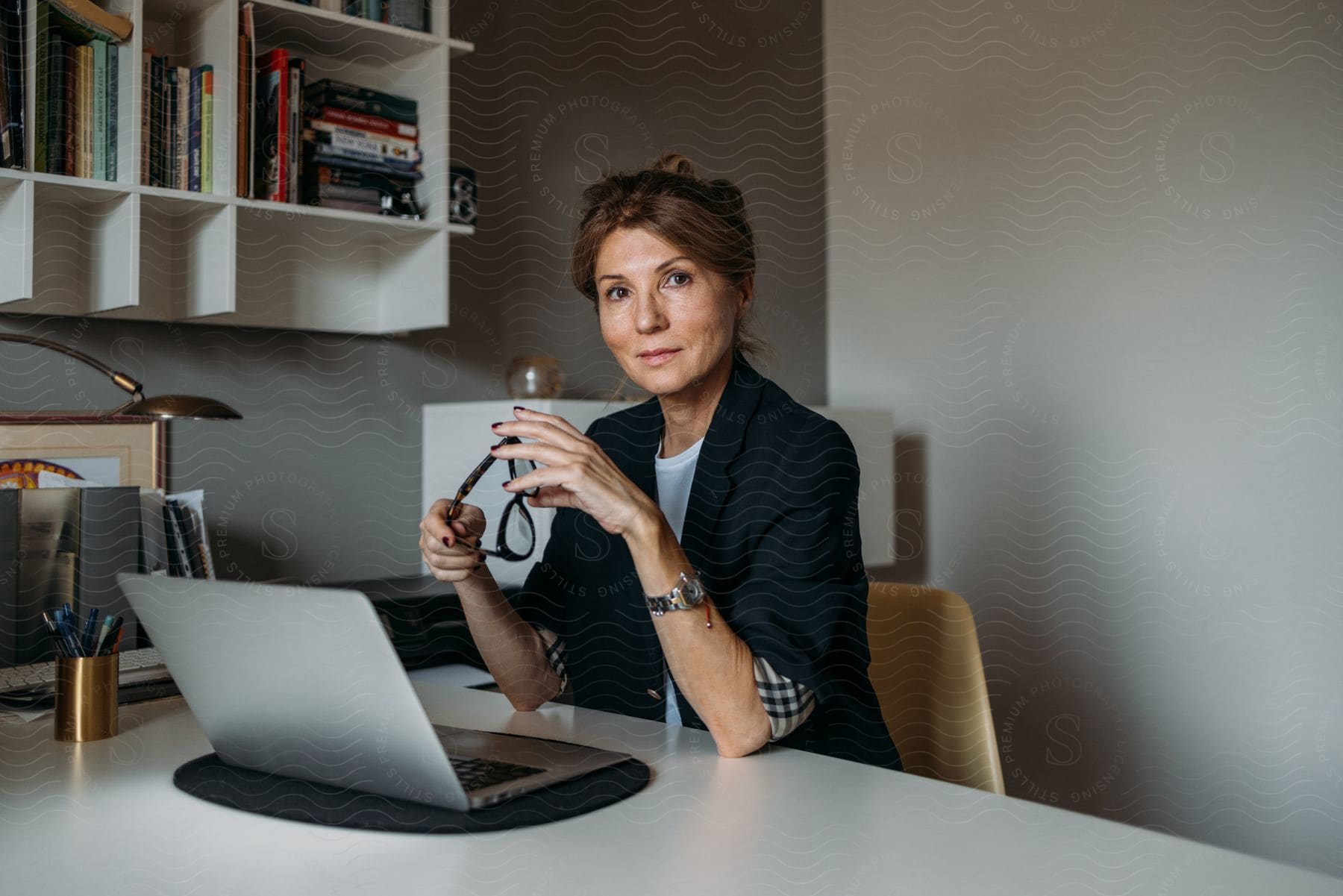 Woman sitting at an office desk holding a pair of prescription glasses in front of her laptop.