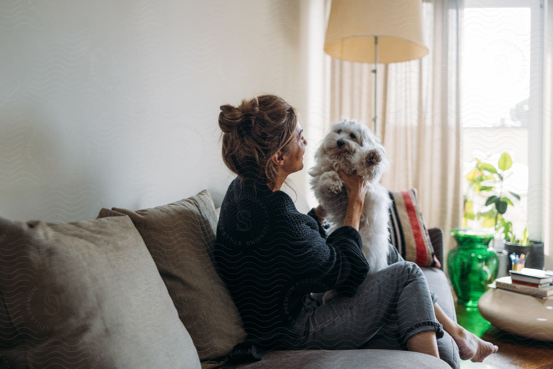 A woman, dressed in a black cardigan and jeans, sits on a couch, cradling a Maltese dog in her arms in the living room