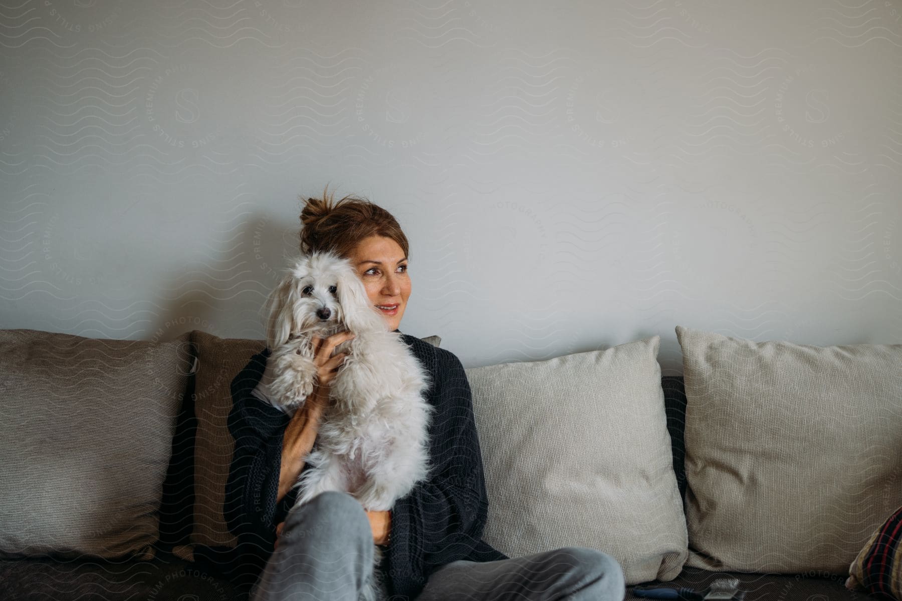 a woman with brown hair sits on a couch, dressed in a black cardigan and jeans, holding a white dog and smiling in the living room