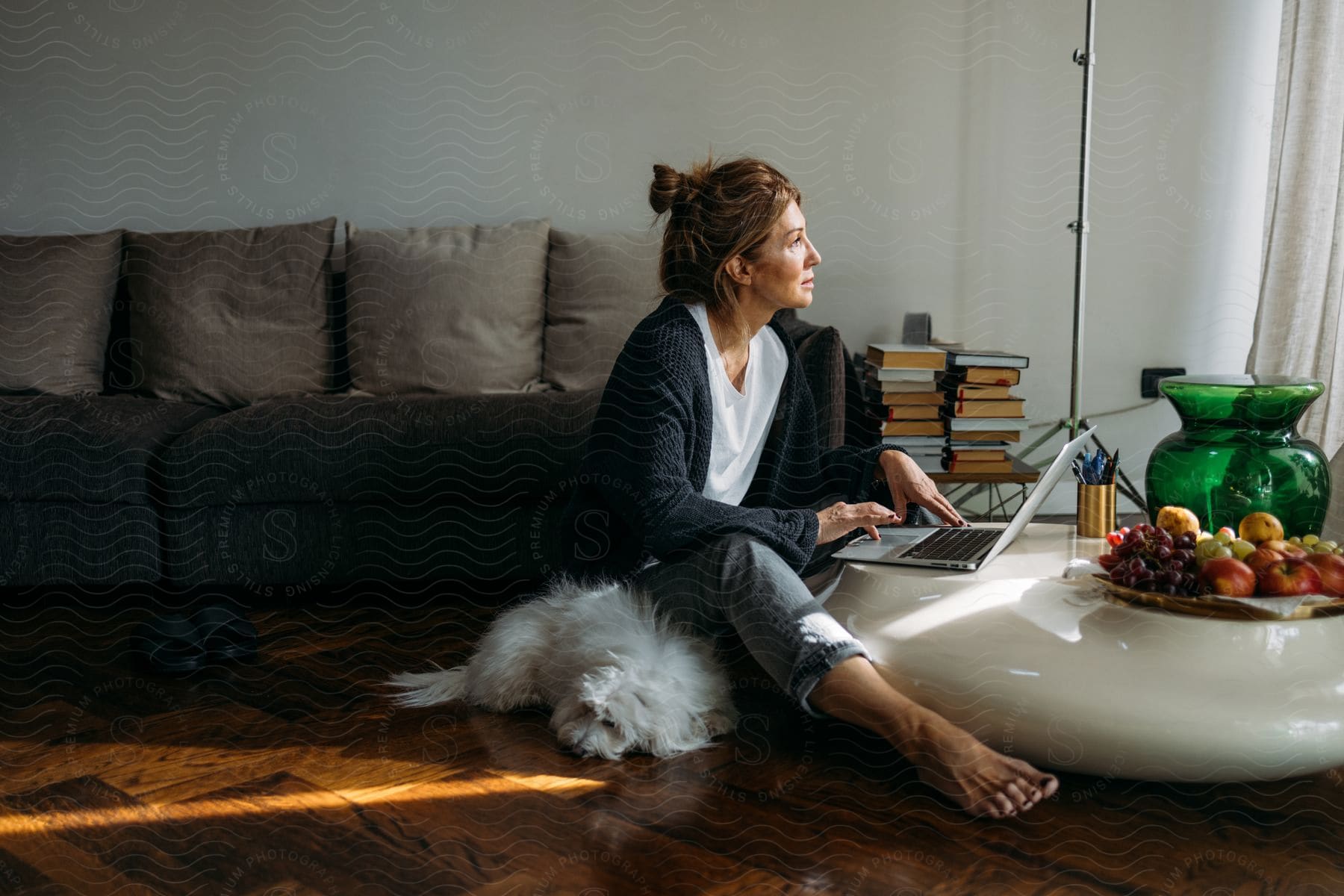 A white woman sits on the floor in her sunlit living room during the day. Her laptop rests on a white round marble table adorned with fresh fruits. A white Lhasa Apso dog lounges nearby, and a couch stands behind her.