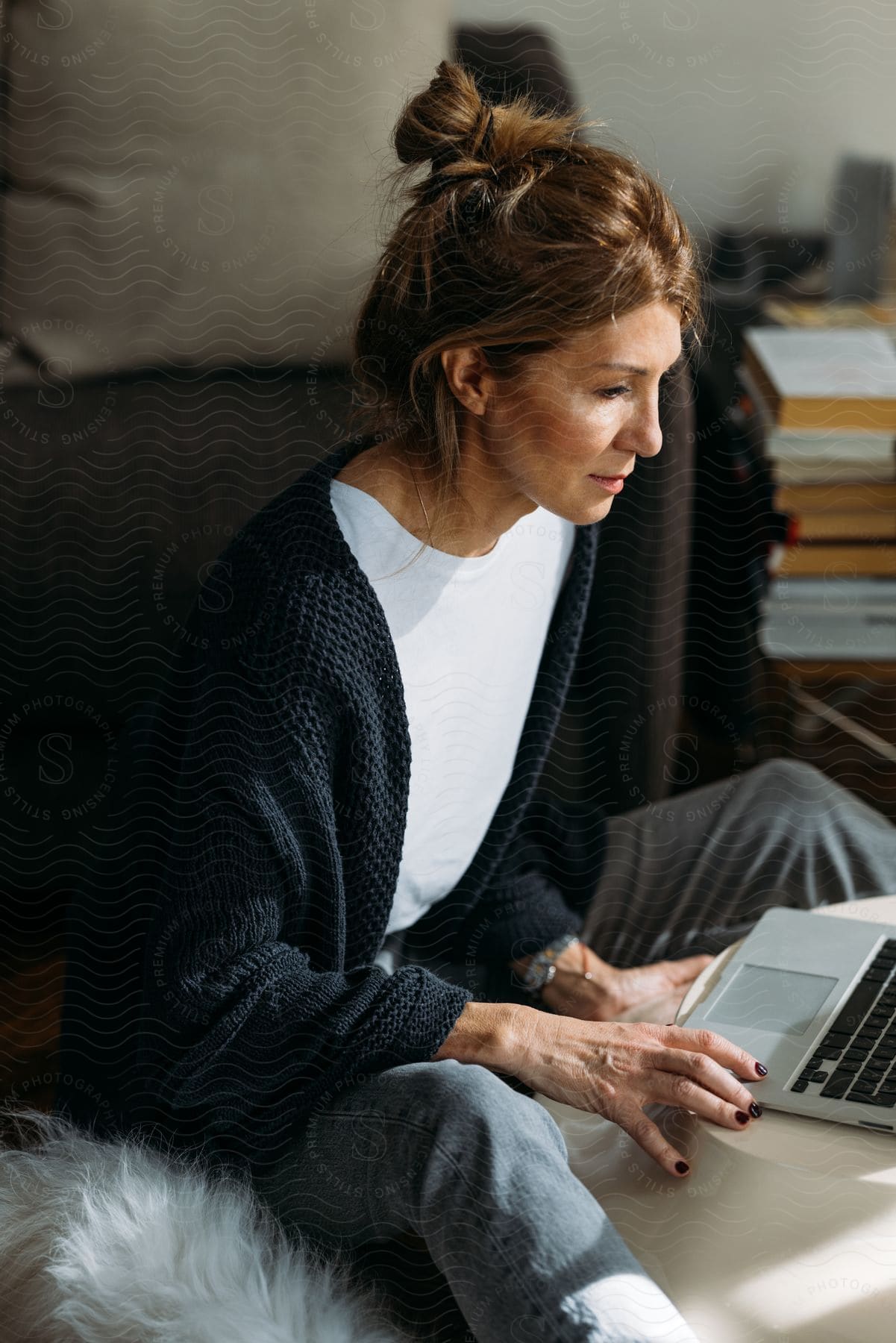 Stock photo of a woman with brown hair is seated on a couch, wearing a black cardigan and jeans, while using a laptop next to a white dog.