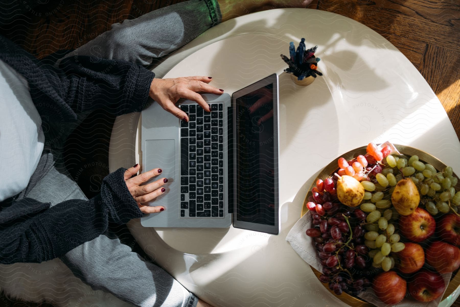 Young Woman Sitting At A Round Table With A Big Bowl Of Fruit And Typing On A Laptop Computer