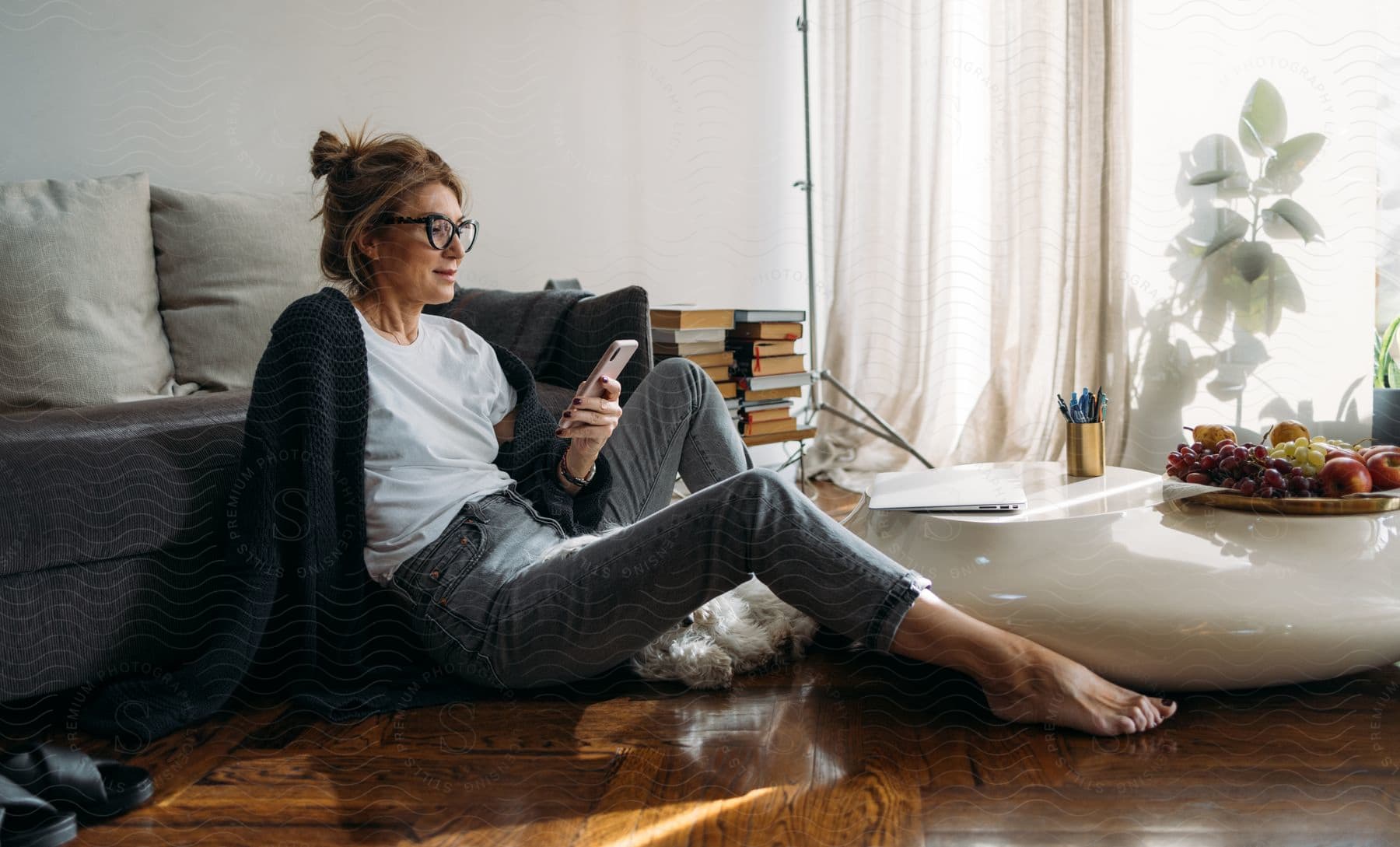 A woman in grey pants and a black sweater checks her phone while sitting on the floor.