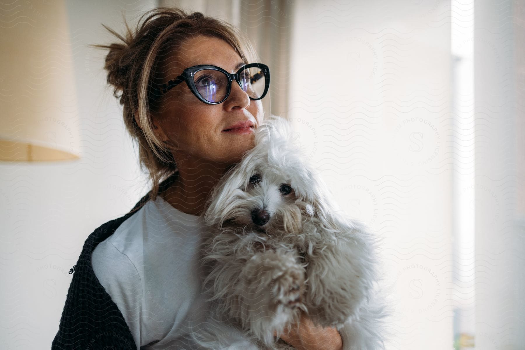 Woman in glasses embracing white dog indoors, looking to the side.