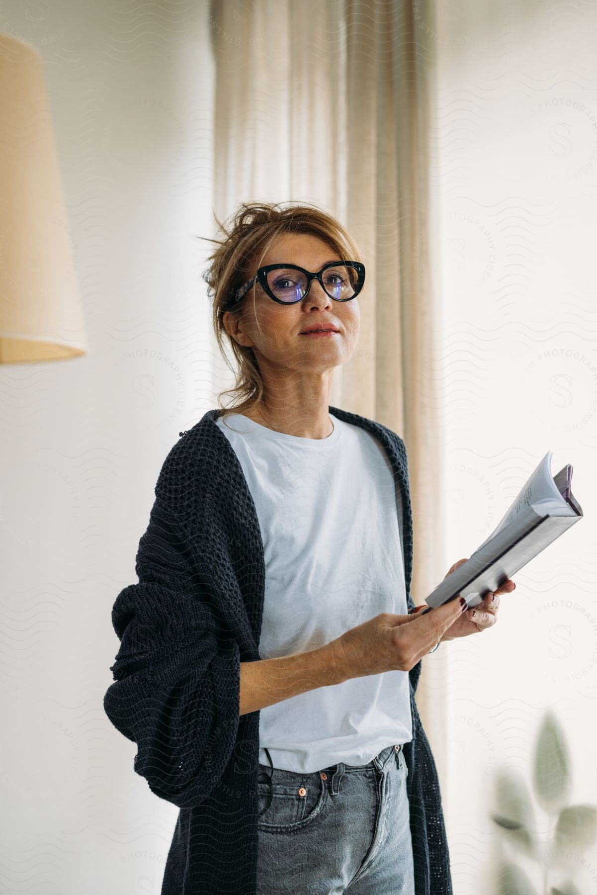 A smiling woman wearing stylish glasses stands indoors while holding a open book.