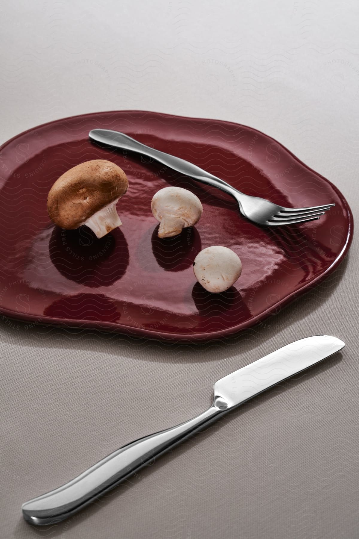 Three mushrooms on a red plate and there is a fork inside the plate and outside there is a knife on a white surface