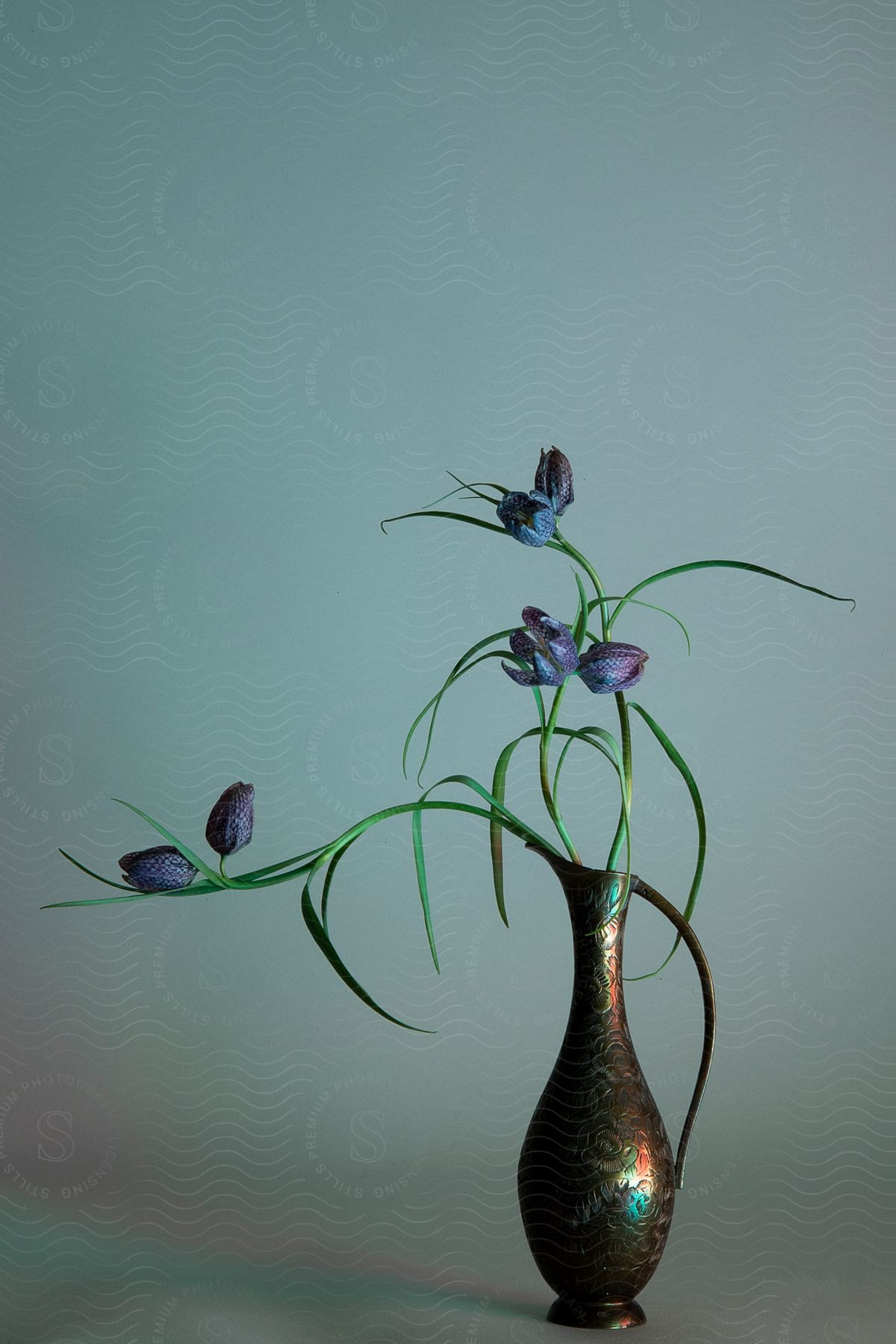 Dramatic flower arrangement of several purple flowers in a copper vase with a long, slender handle.