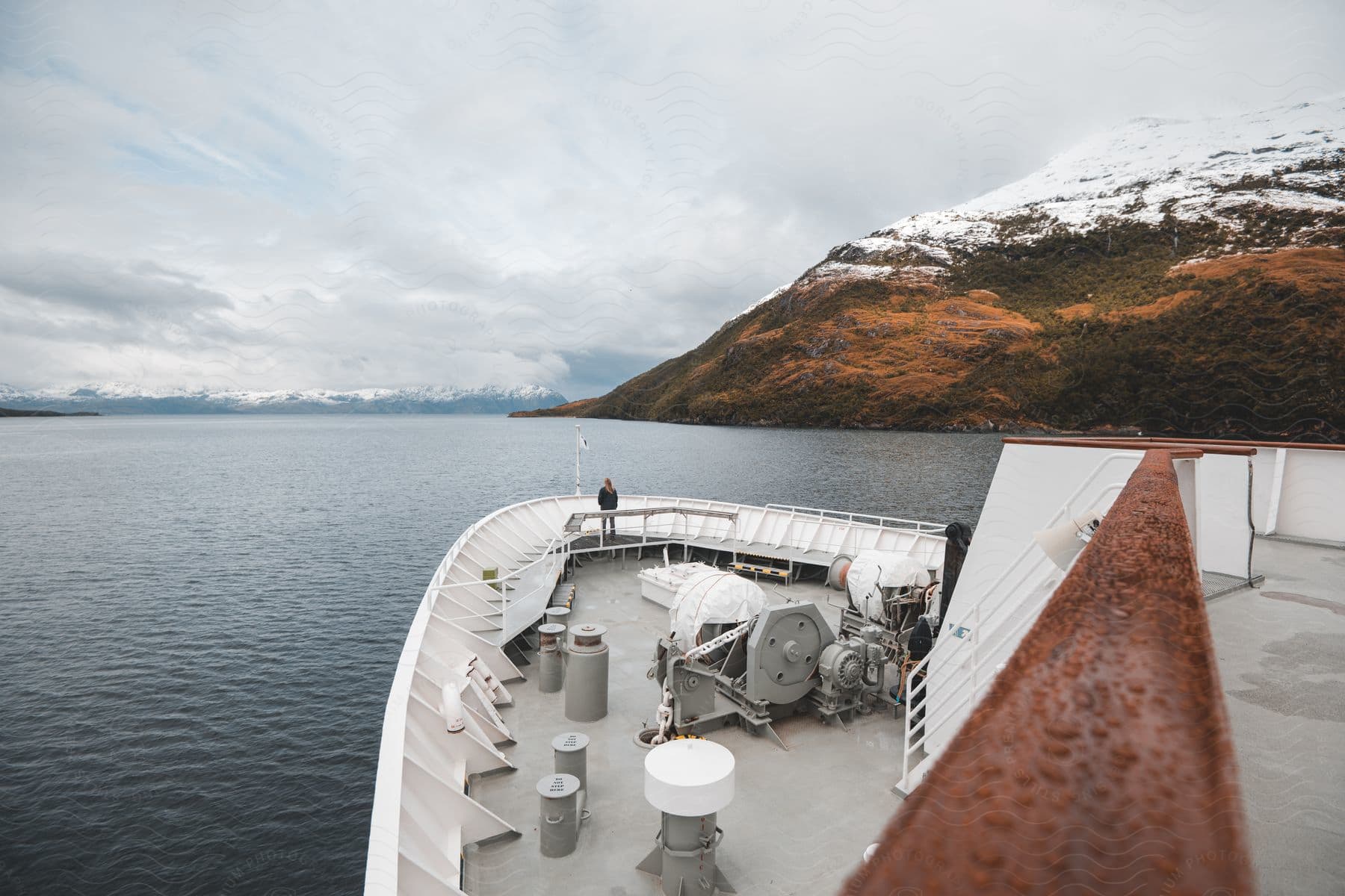 Distant person standing on the deck of a cruise ship, looking out at a scenic fjord with steep cliffs on a cloudy day