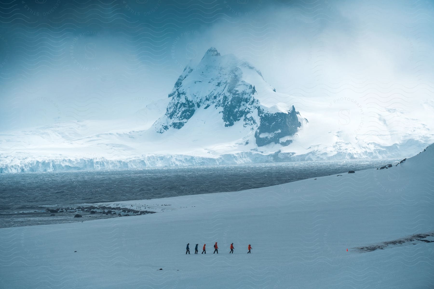 People hike across a snowy field with a snow-capped mountain and iceberg near the Arctic Ocean.