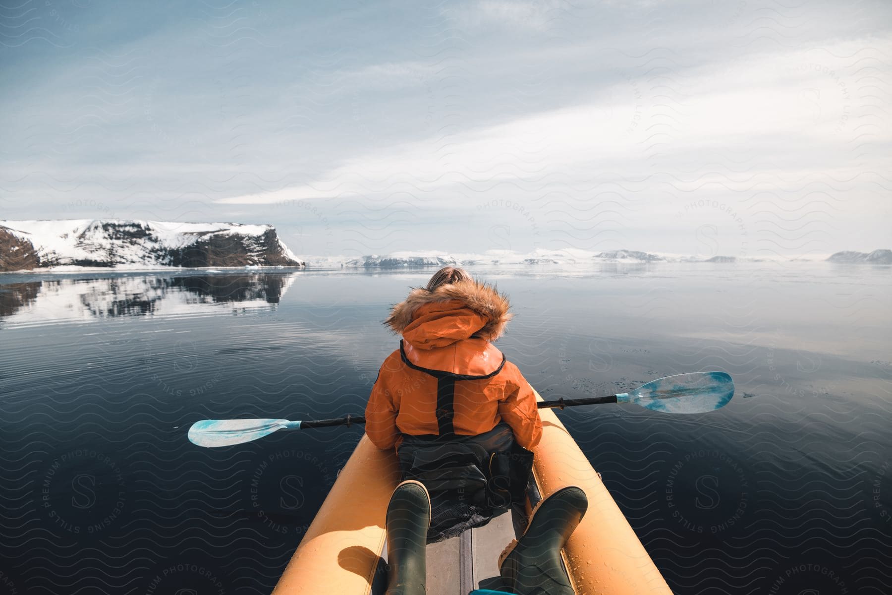 A person is seated at the front of a canoe, clad in a parka, gripping a double-bladed paddle, with another individual in boots positioned behind them, amidst the icy waters of the Arctic Sea with snow filled rocked in the background