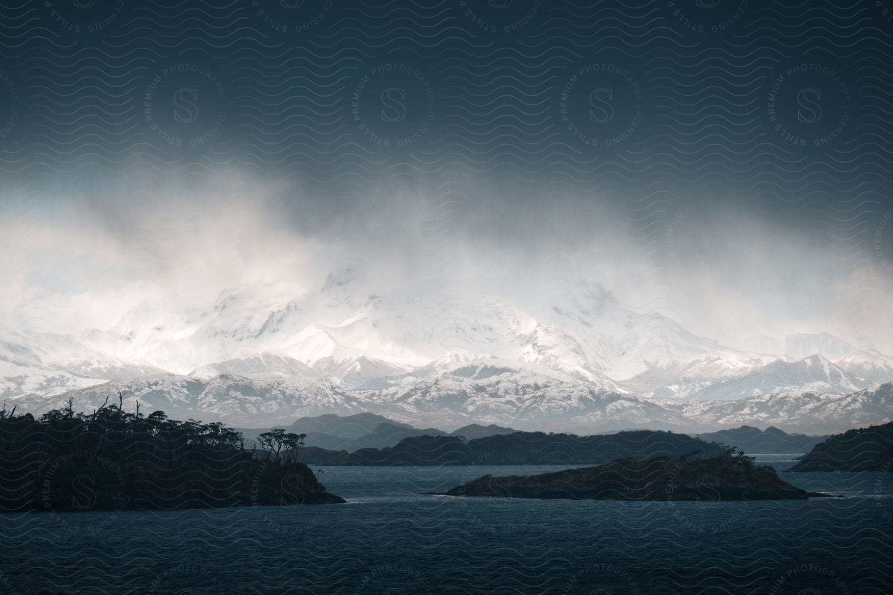 A dense cloud obscures the sky above a river, which is encircled by small, forested islands. In the distance, a mountain stands with its summit bathed in sunlight and capped with snow.