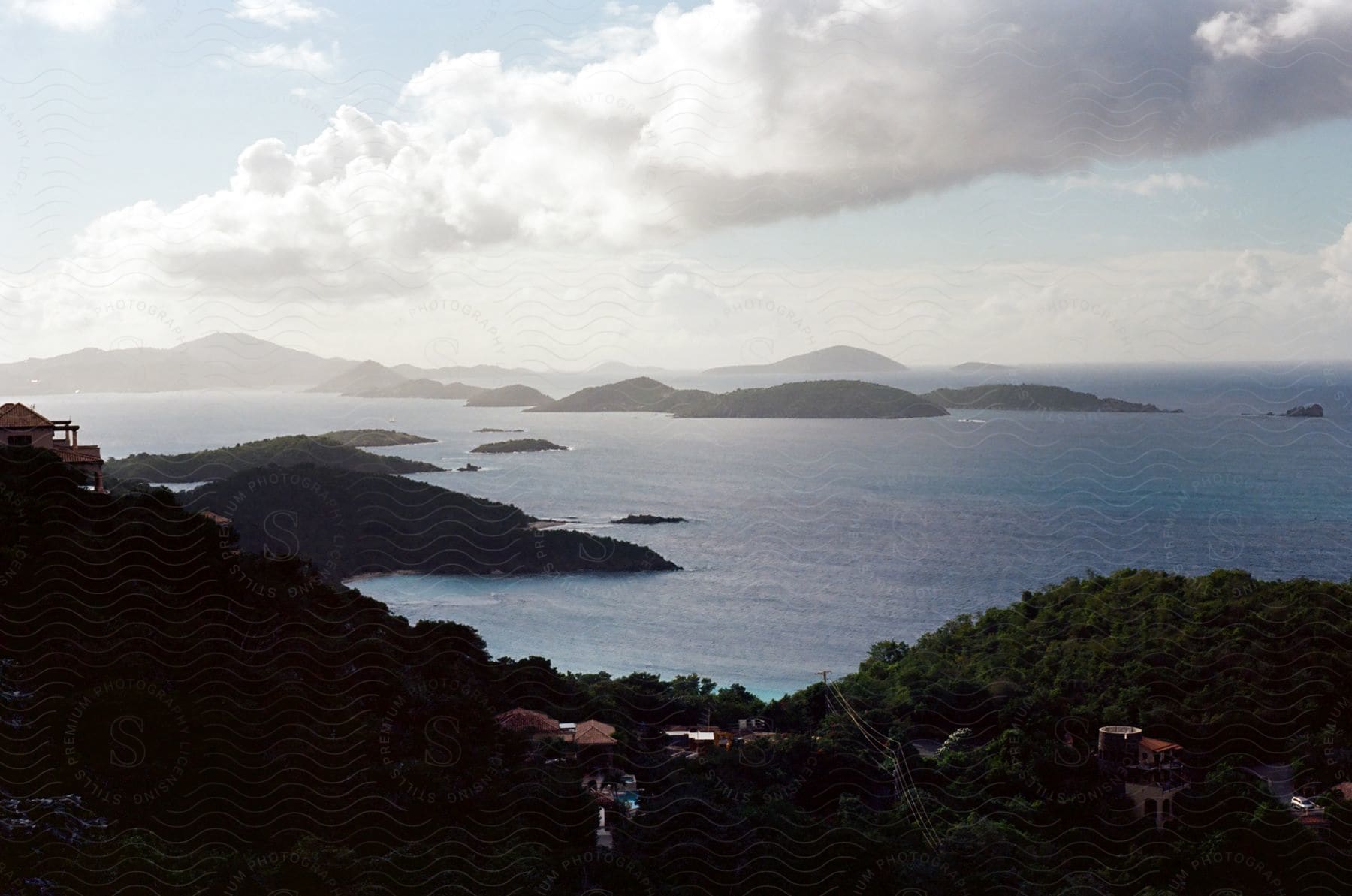 A serene view of a coastal landscape with lush vegetation, calm waters and distant islands under a partly cloudy sky.