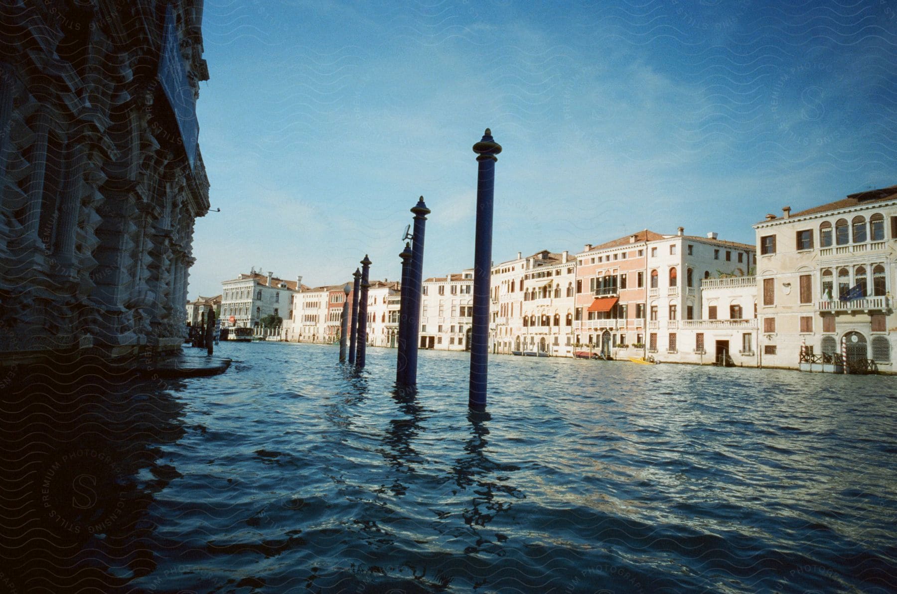 A serene scene of a Venetian canal with historic buildings and mooring posts. The blue sky reflects off the calm water, while the buildings show off Venice's classic architecture.