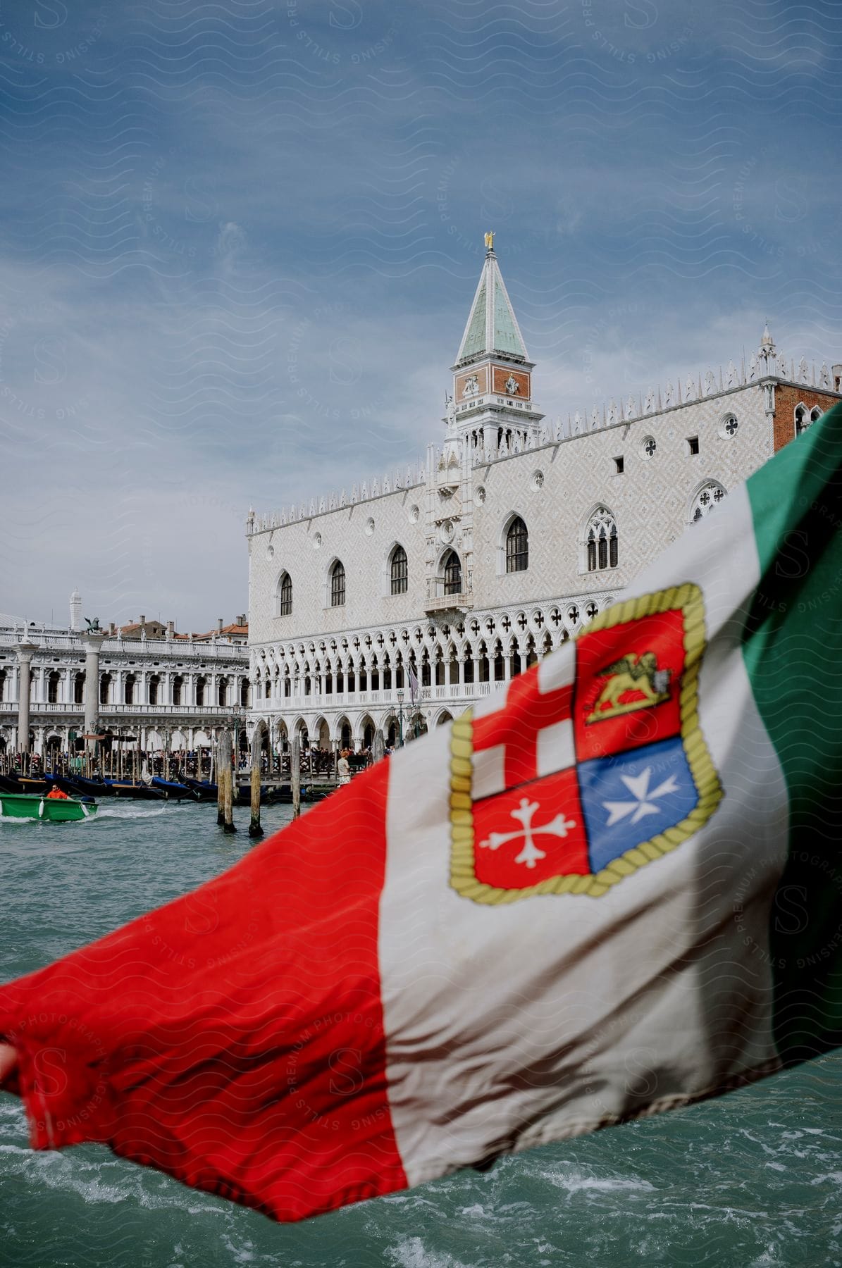 An Italian flag with a coat of arms waves in the foreground, partially obscuring the view of Doge's Palace, a landmark of Venice with ornate Gothic architecture, seen from the water on a sunny day.